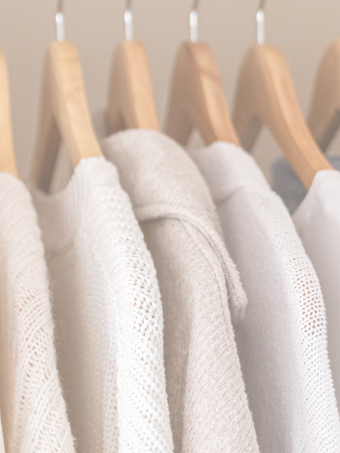 A row of beige, white, and off-white knitted sweaters hanging on wooden hangers on a clothing rack.