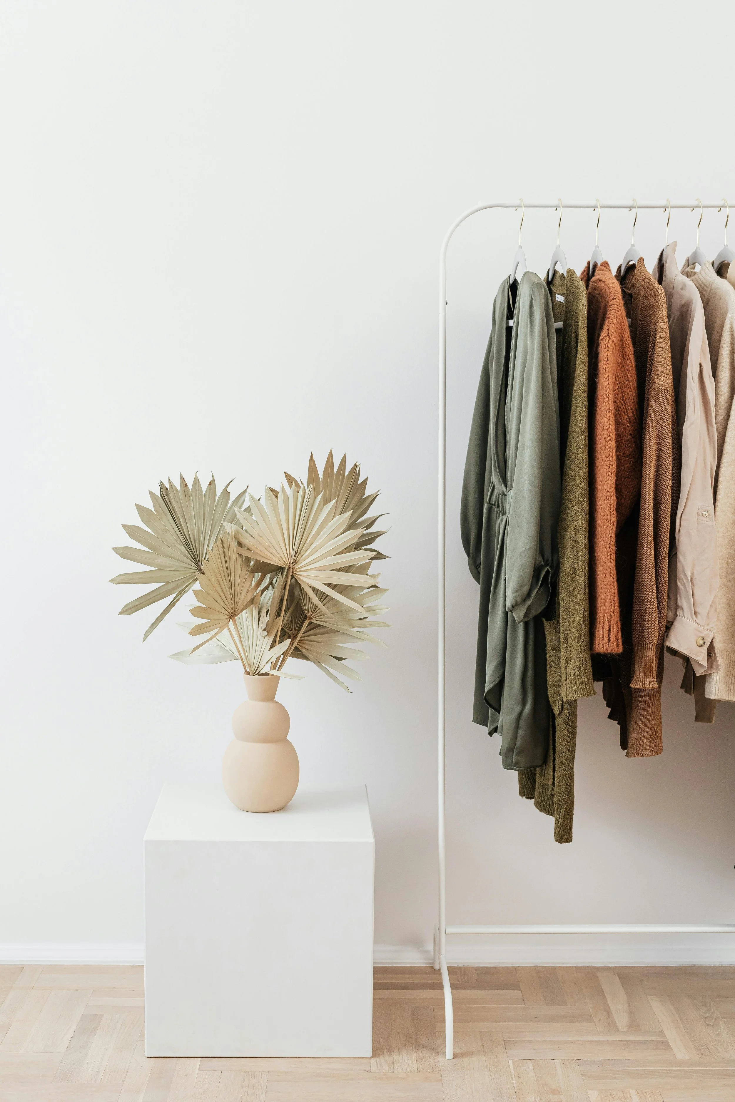 A clothing rack with various sweaters and jackets hanging next to a beige decorative vase with dried palm leaves inside, placed on a white pedestal against a plain white wall.