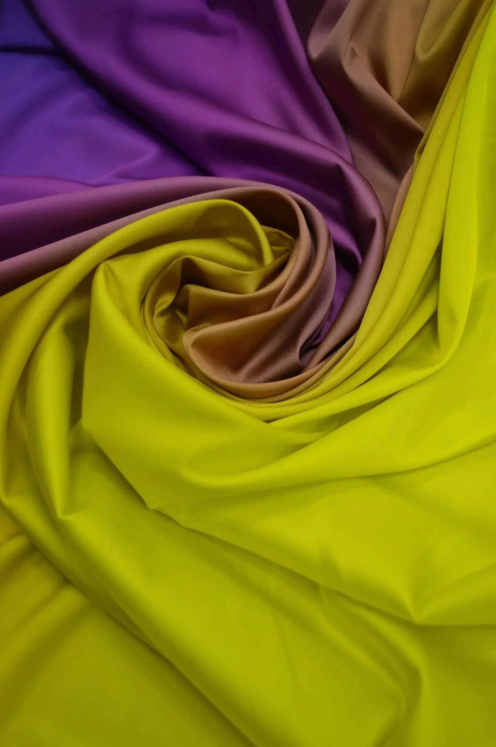 Close-up photograph of colorful satin fabric in purple, brown, and yellow, artistically arranged with folds and a swirling pattern.