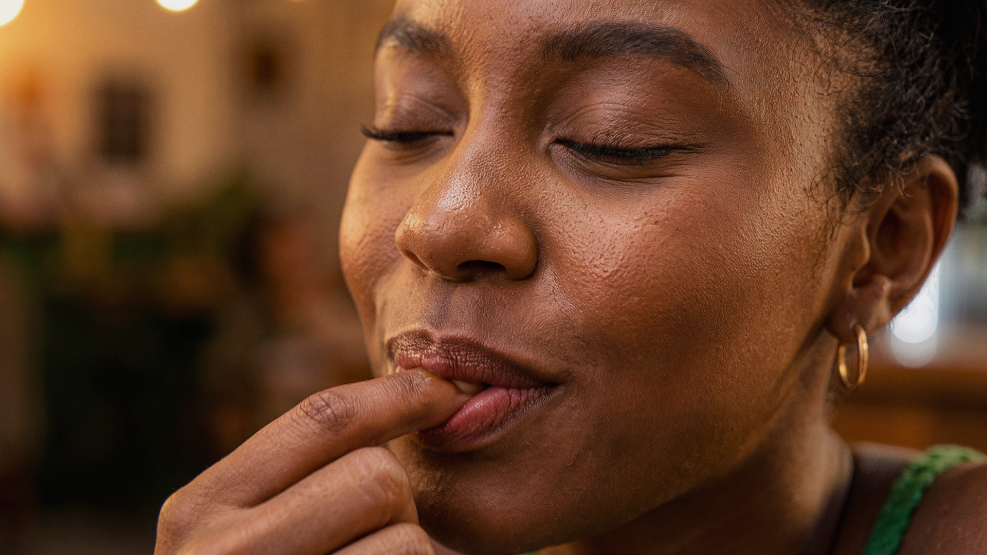 Close-up de uma mulher com pele morena, olhos fechados, mostrando detalhes de sua pele e sobrancelhas, tocando os lábios com o dedo.