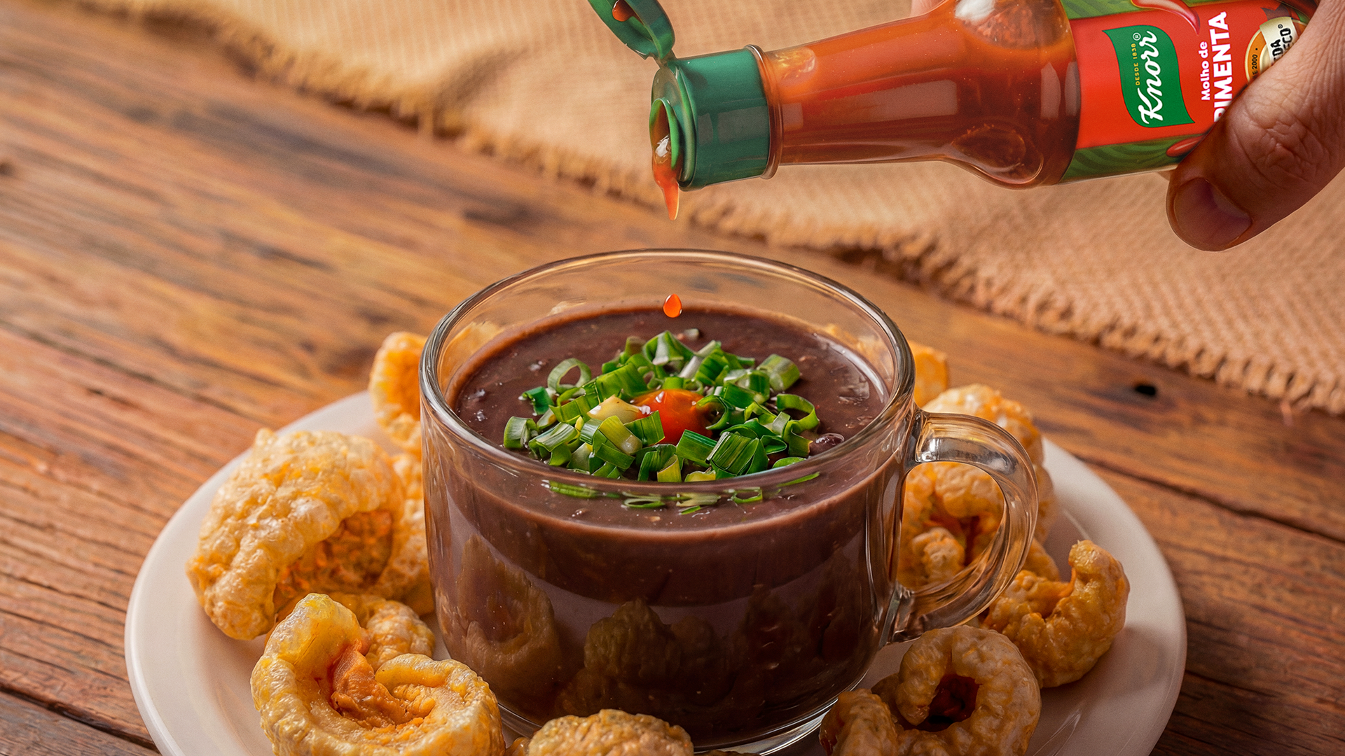 Molho de pimenta sendo colocado em um copo de vidro com sopa preta, decorada com cebolinha e acompanhada de pedaços de torresmo em uma tigela branca sobre uma mesa de madeira.
