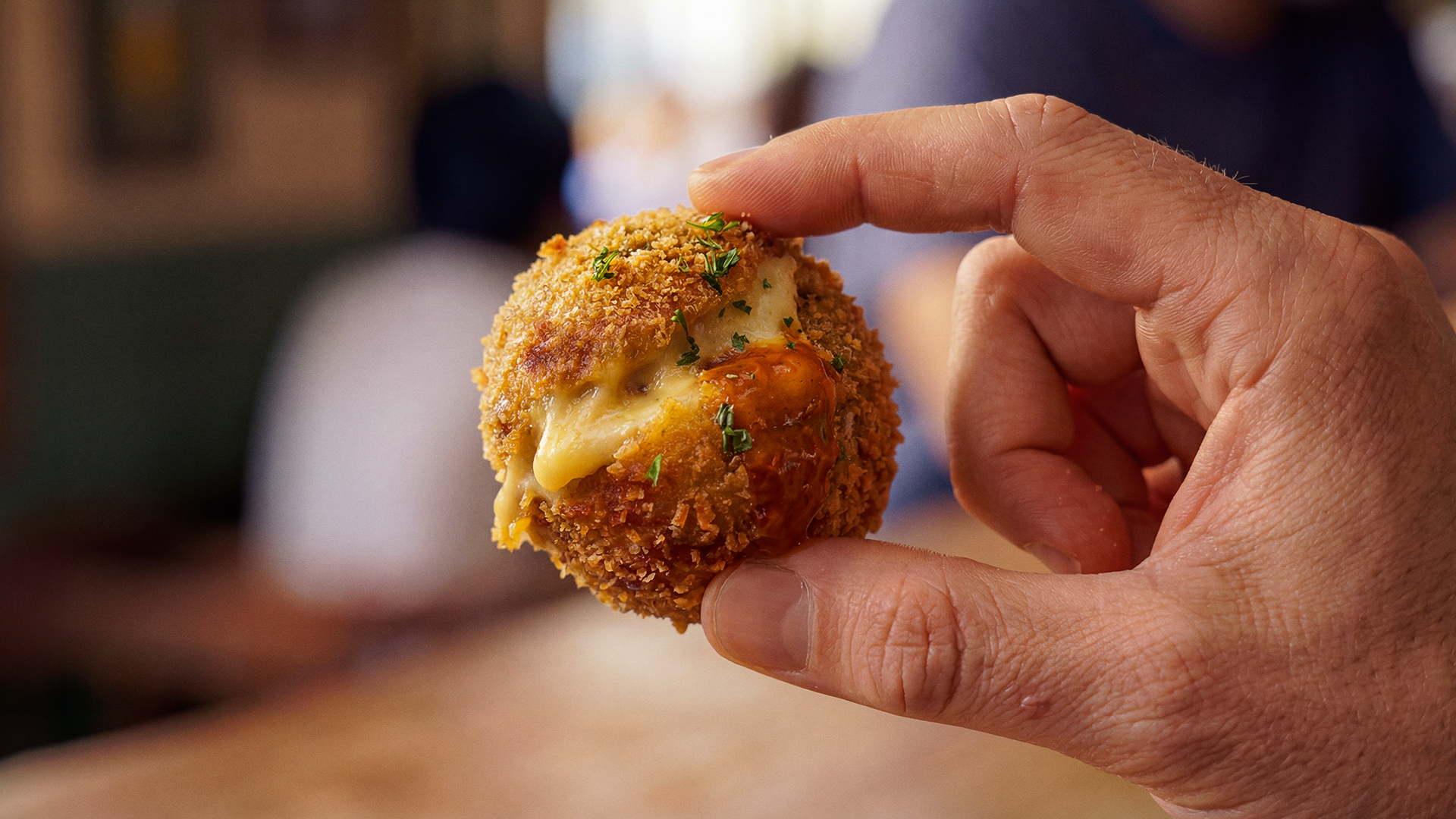Pessoa segurando uma croquete de queijo empanada, com molho e ervas por cima.