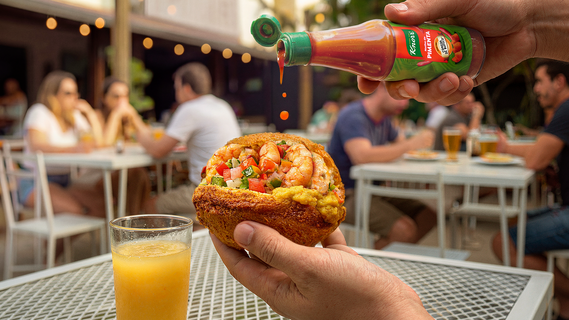 Mão segurando uma taça de suco de laranja, enquanto outra pessoa segura uma porção de bolinho recheado com camarão, acompanhada de molho apimentado. Em fundo, pessoas conversando em uma área de restaurante ao ar livre.