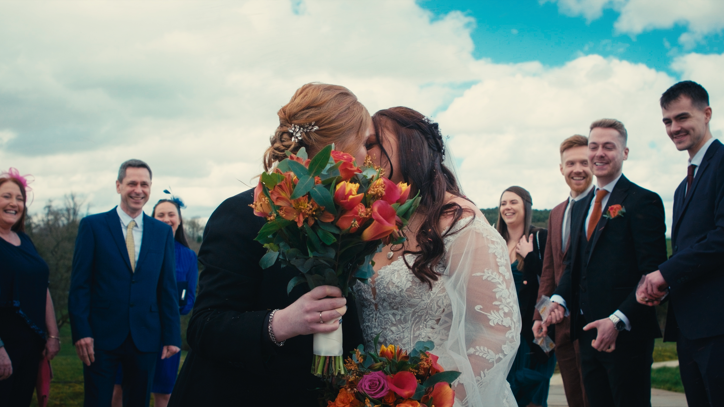 Two women, one with red hair and the other with dark hair, kissing in wedding attire, holding a bouquet of colorful flowers, surrounded by friends in formal wear outdoors on a cloudy day.