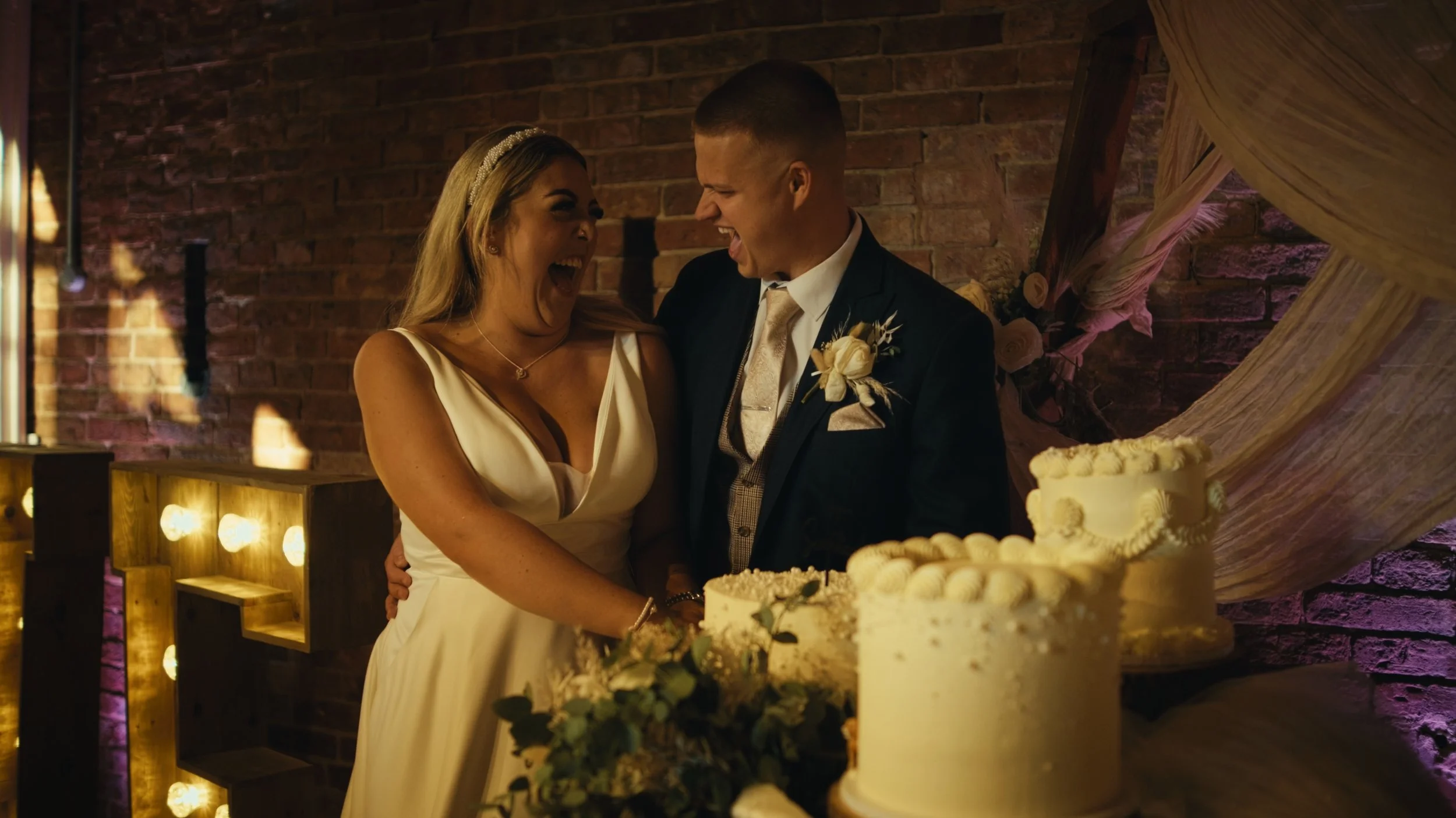 A bride and groom smiling and celebrating at their wedding reception, standing near a wedding cake, with a brick wall in the background.