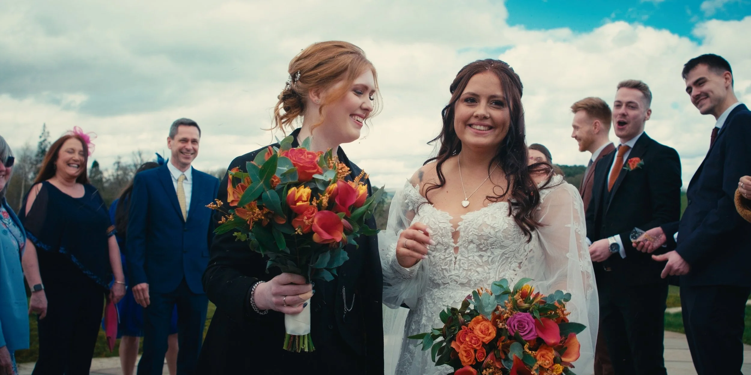 A wedding celebration outdoors with two smiling women holding bouquets, surrounded by friends dressed in formal attire under a cloudy sky.