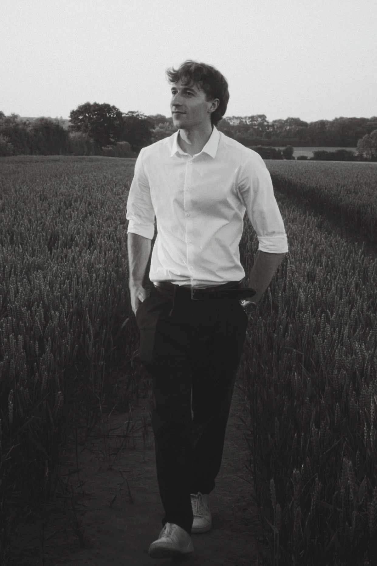 A young man with wavy hair wearing a white shirt, black pants, and white sneakers walking through a wheat field on a clear day.