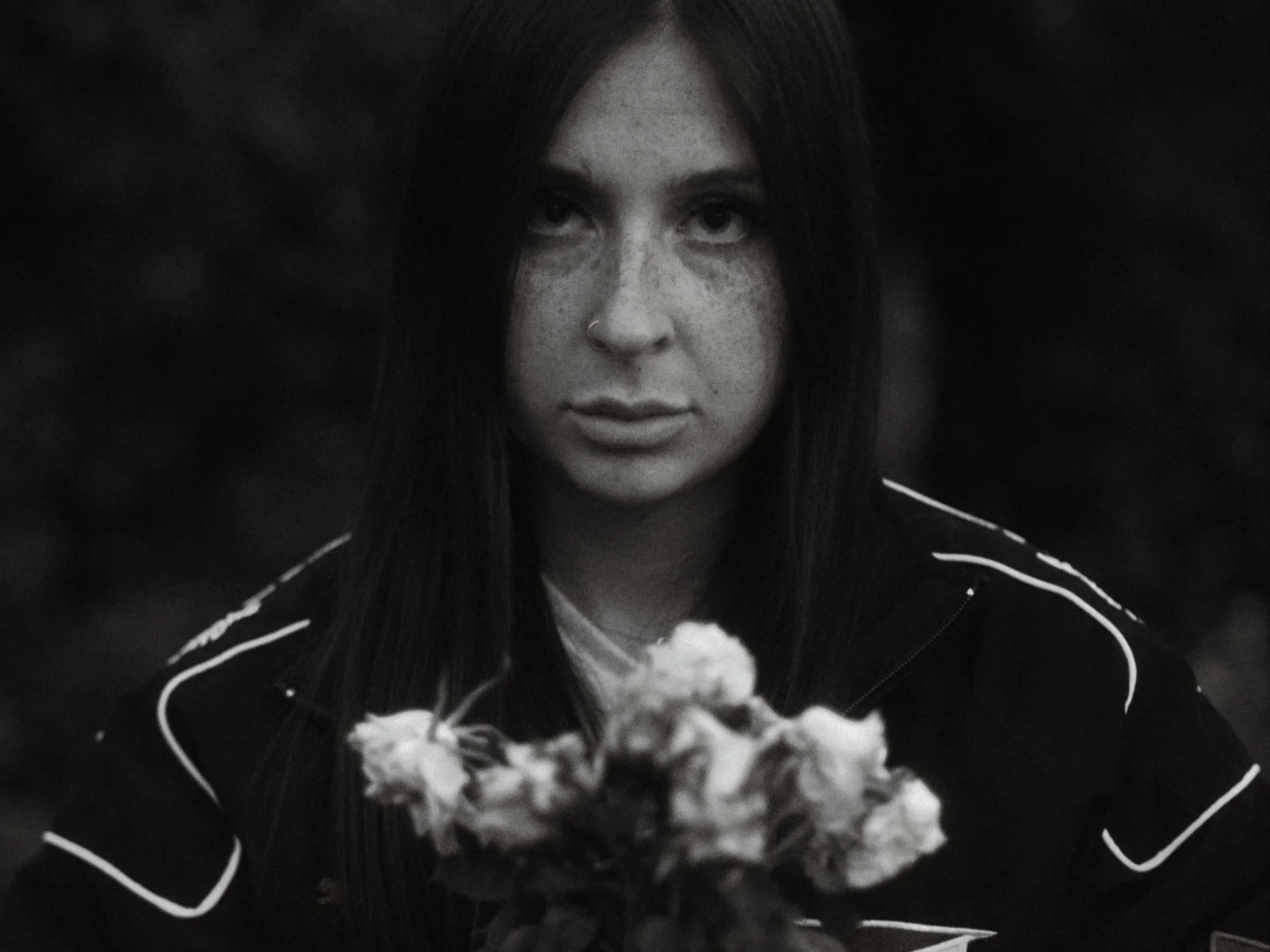 A young woman with dark hair, freckles, and a nose piercing looking directly at the camera with a serious expression, wearing a dark jacket with white piping, holding a flower.