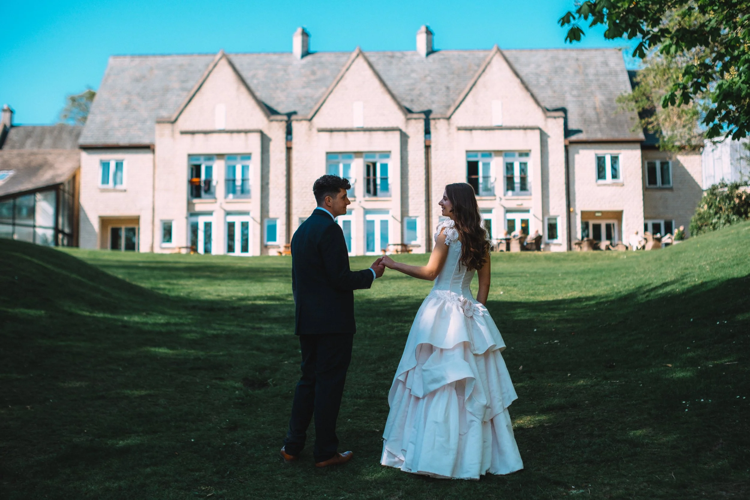 A bride and groom holding hands and smiling at each other outdoors in front of a large house with multiple windows, surrounded by trees and greenery.