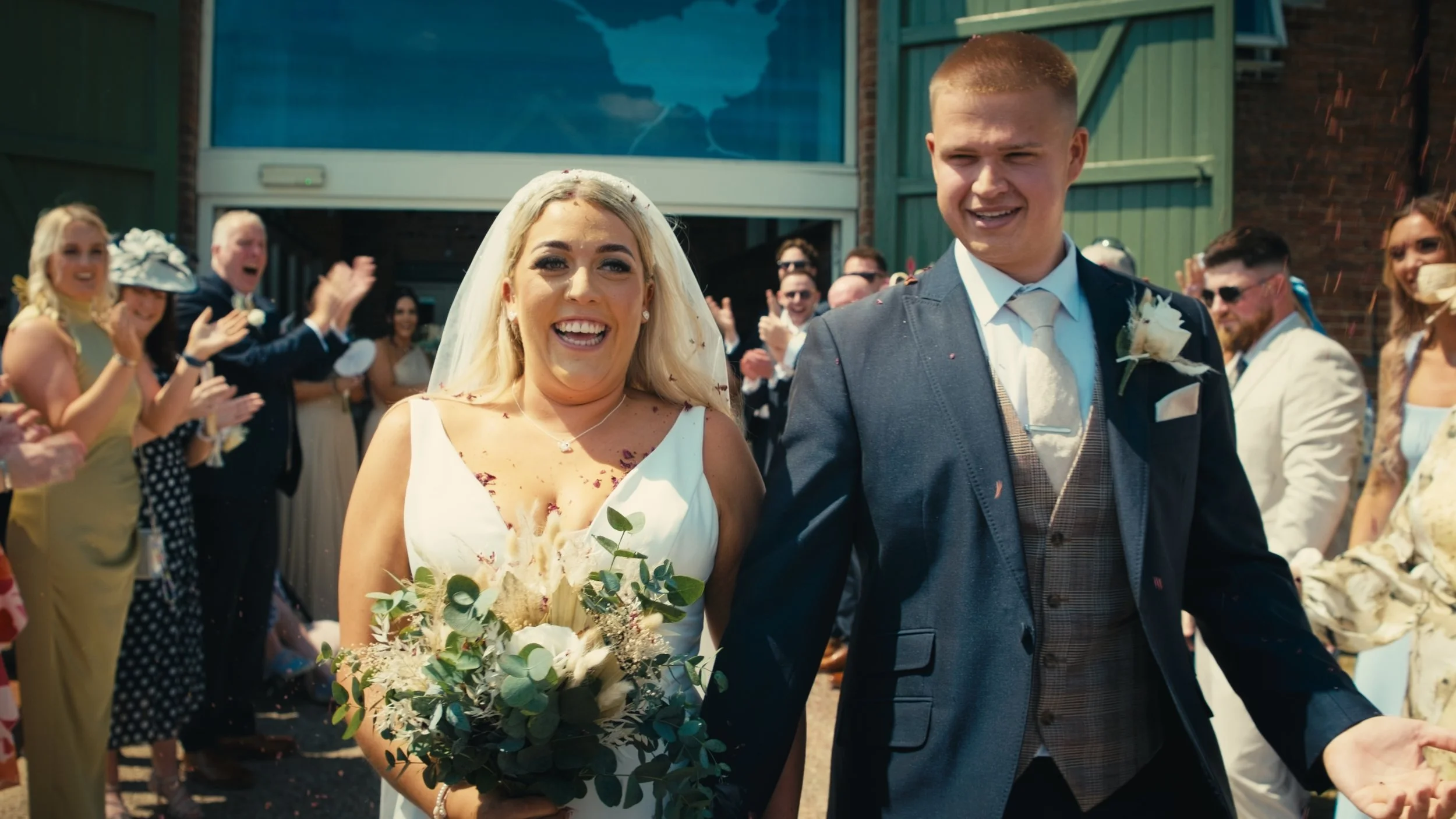 A bride and groom walking together at their wedding celebration, surrounded by smiling guests clapping outdoors.