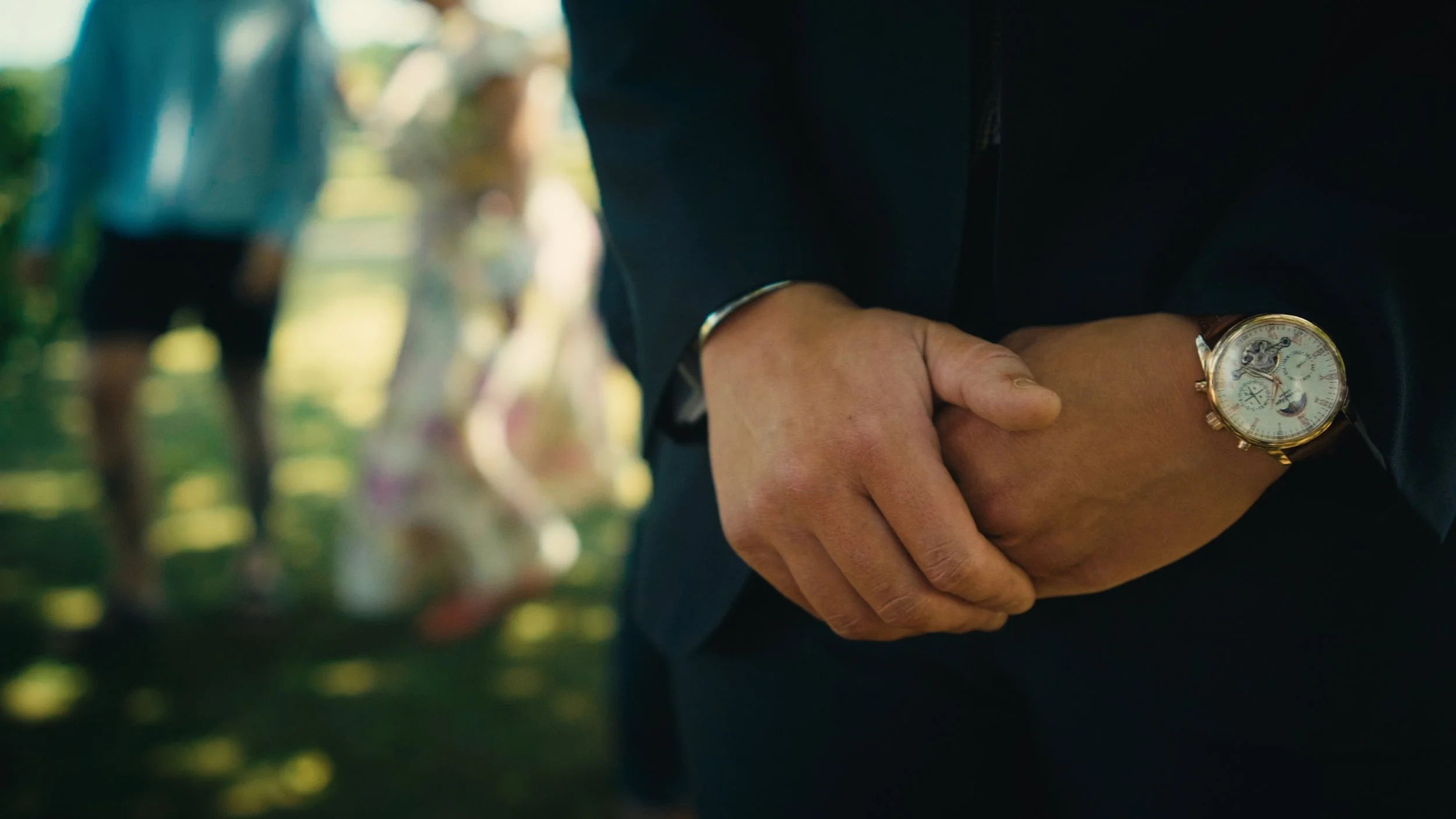 Close-up of a person in a suit with their hands clasped, wearing a gold watch, at an outdoor gathering with people in the background.