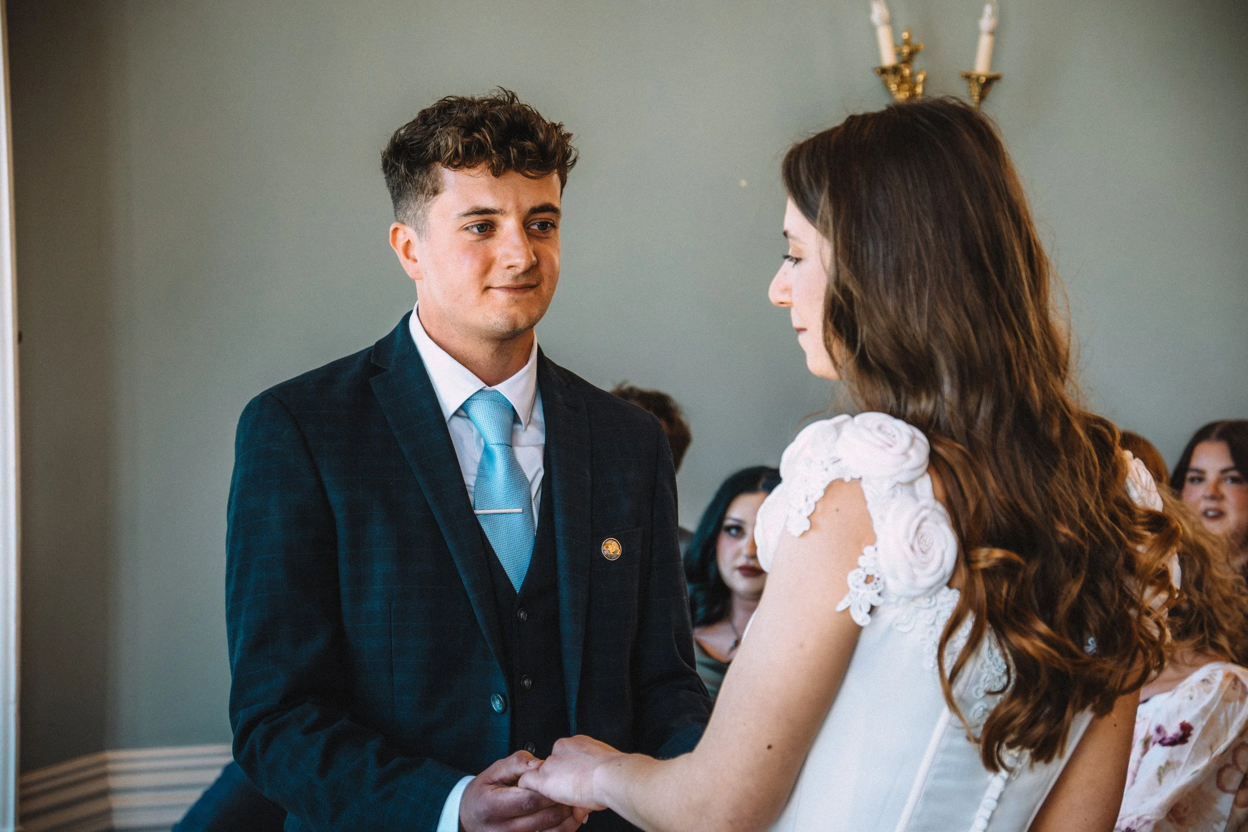 A groom and bride holding hands and exchanging vows during their wedding ceremony, with several guests observing in the background.