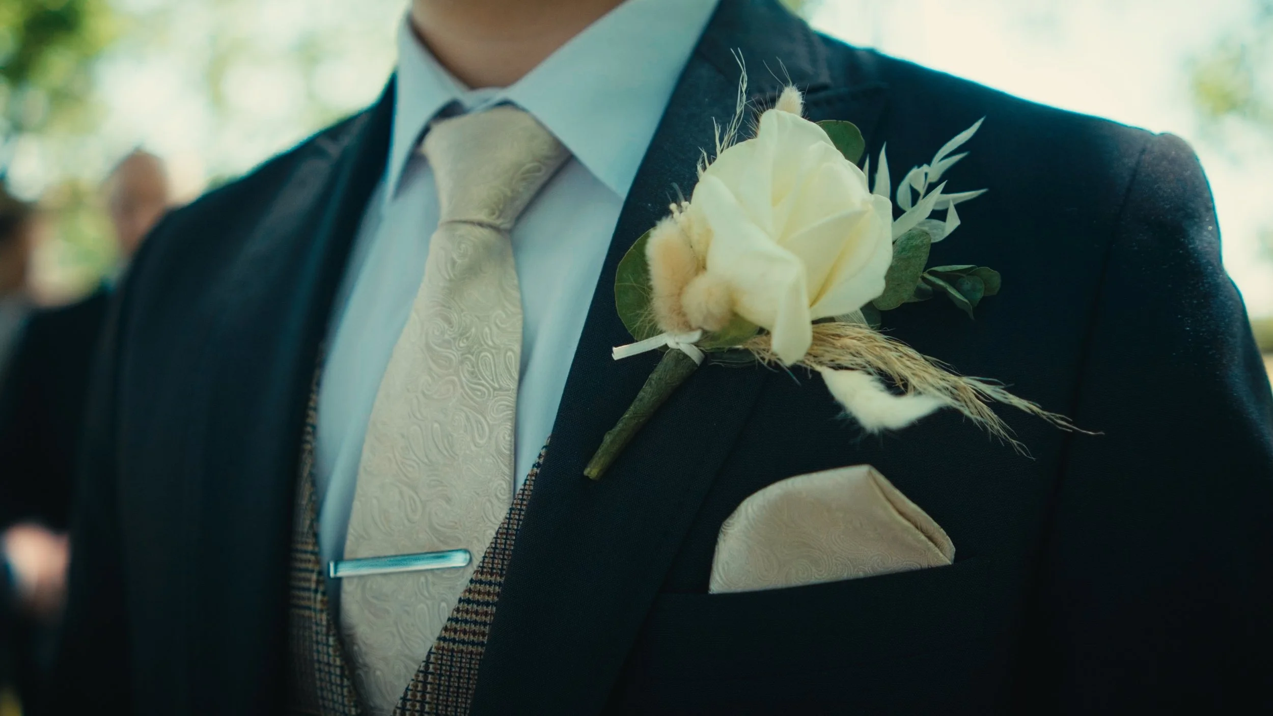 Close-up of a groom's suit with a cream-colored flower boutonniere on his left lapel, wearing a dark suit, a light blue shirt, and a cream-colored patterned tie, outdoors with blurred greenery in the background.