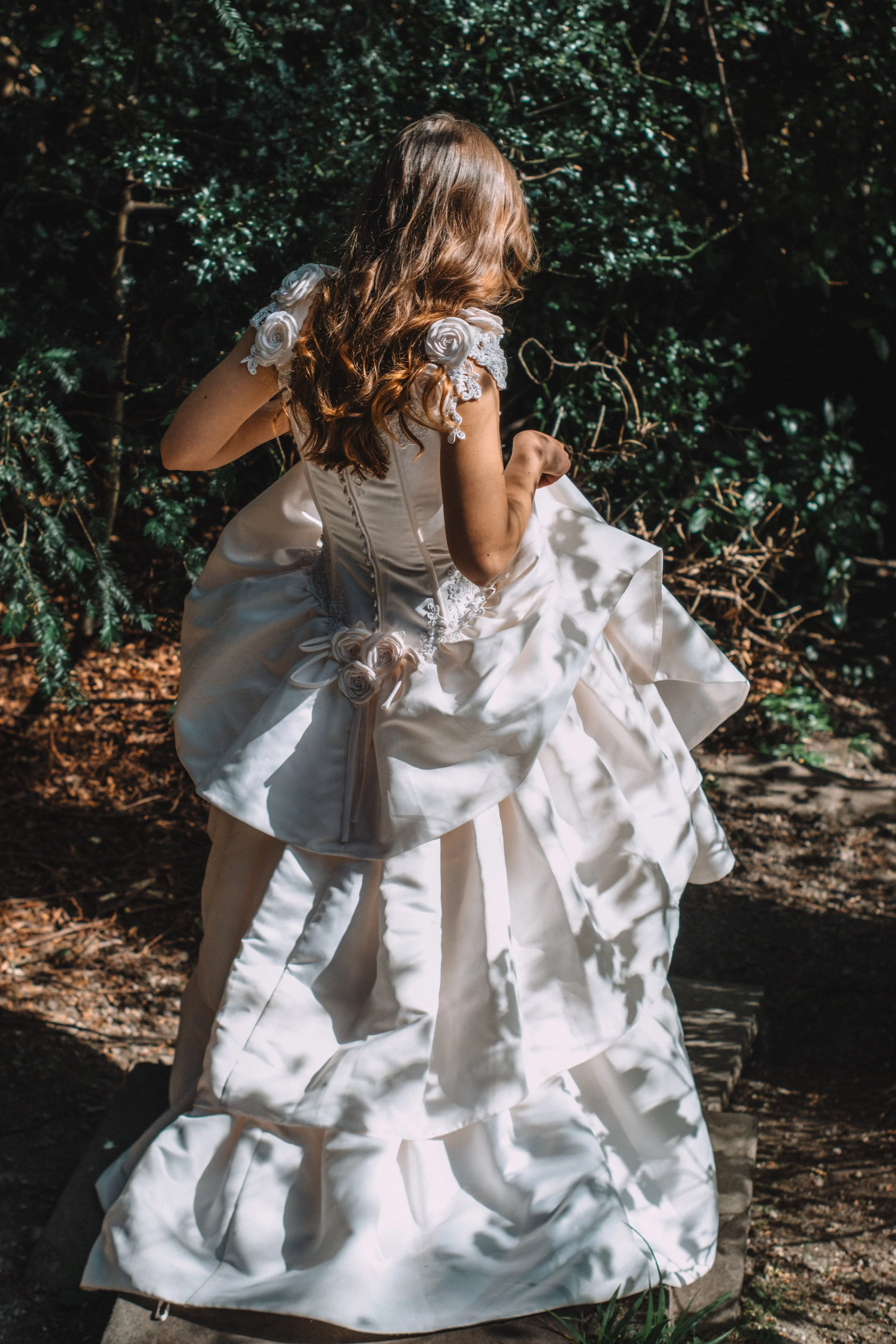 A woman in a white dress with rose embellishments, sitting outdoors in a natural, wooded setting with green foliage.