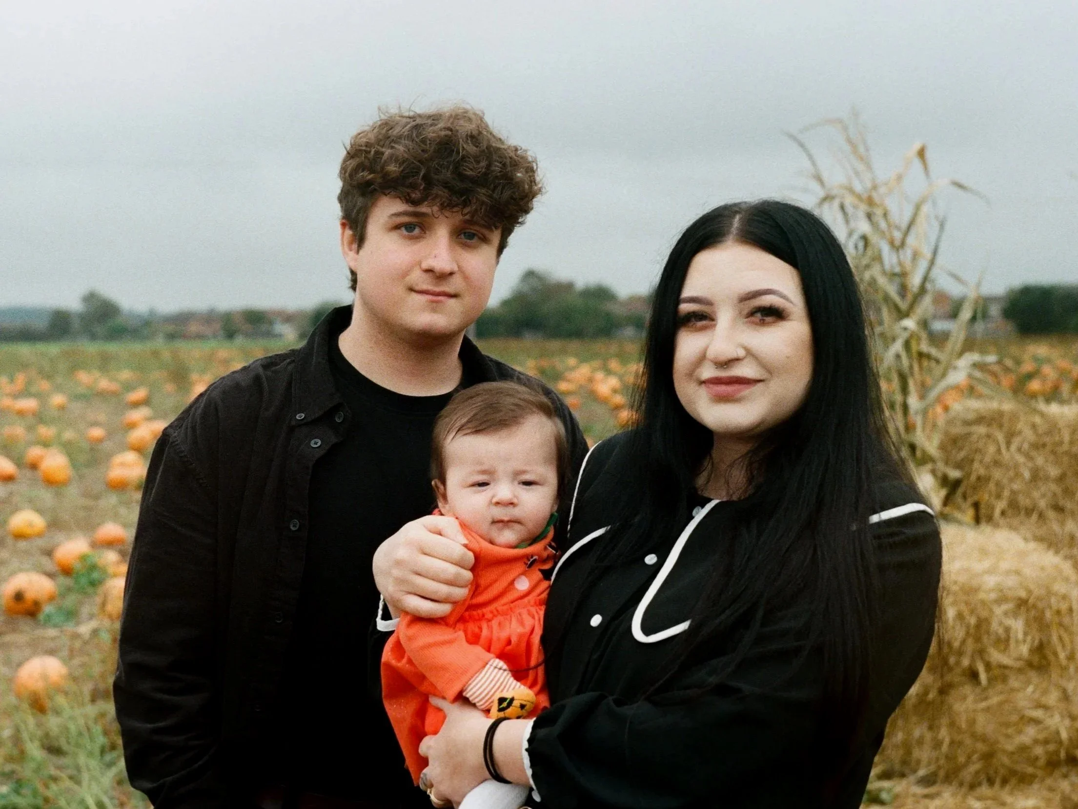 A group of three people outdoors with a pumpkin patch in the background on an overcast day. A young man with curly brown hair, a woman with long black hair and a septum piercing, and a young child with light brown hair, dressed in an orange outfit.