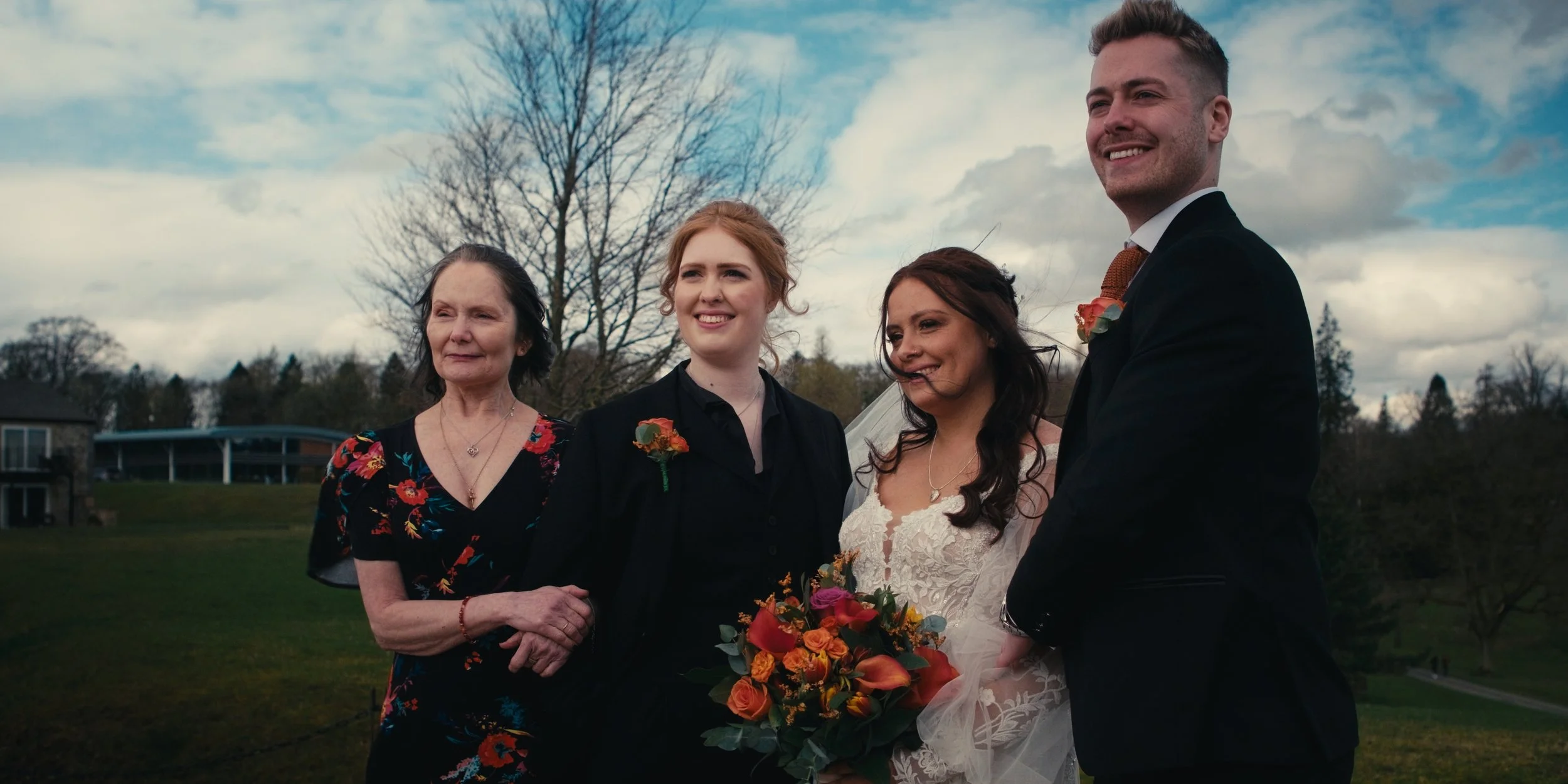 A group of four people standing outdoors on a cloudy day, dressed in formal attire, with the bride holding a bouquet of flowers. They are smiling and looking in different directions.