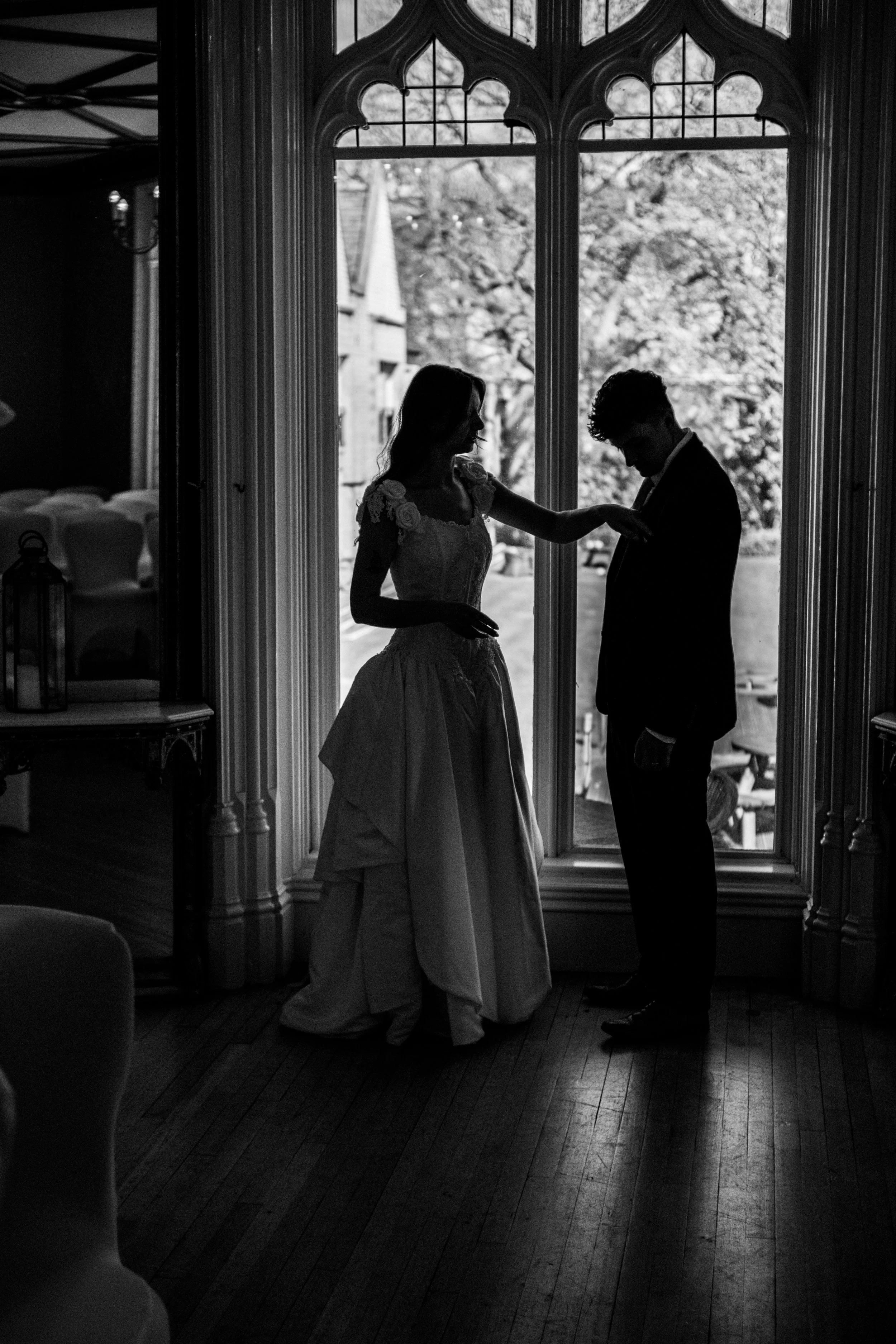 Silhouette of a bride and groom standing inside by a large window, looking at each other, during a wedding or romantic moment.