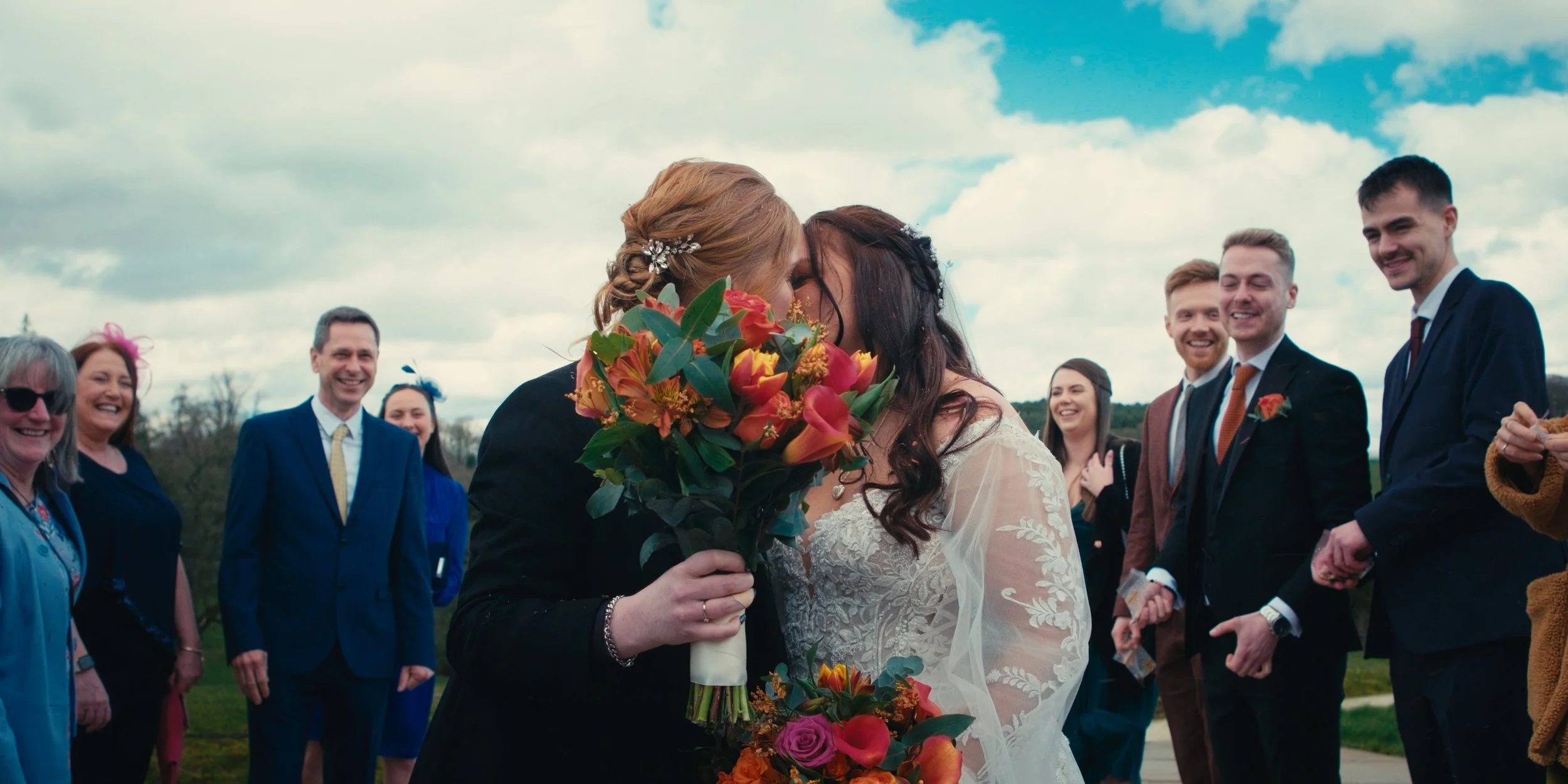 Two women, possibly a bride and groom, are kissing. The woman on the left is holding a colorful bouquet. Several smiling guests are standing around them outdoors on a cloudy day.