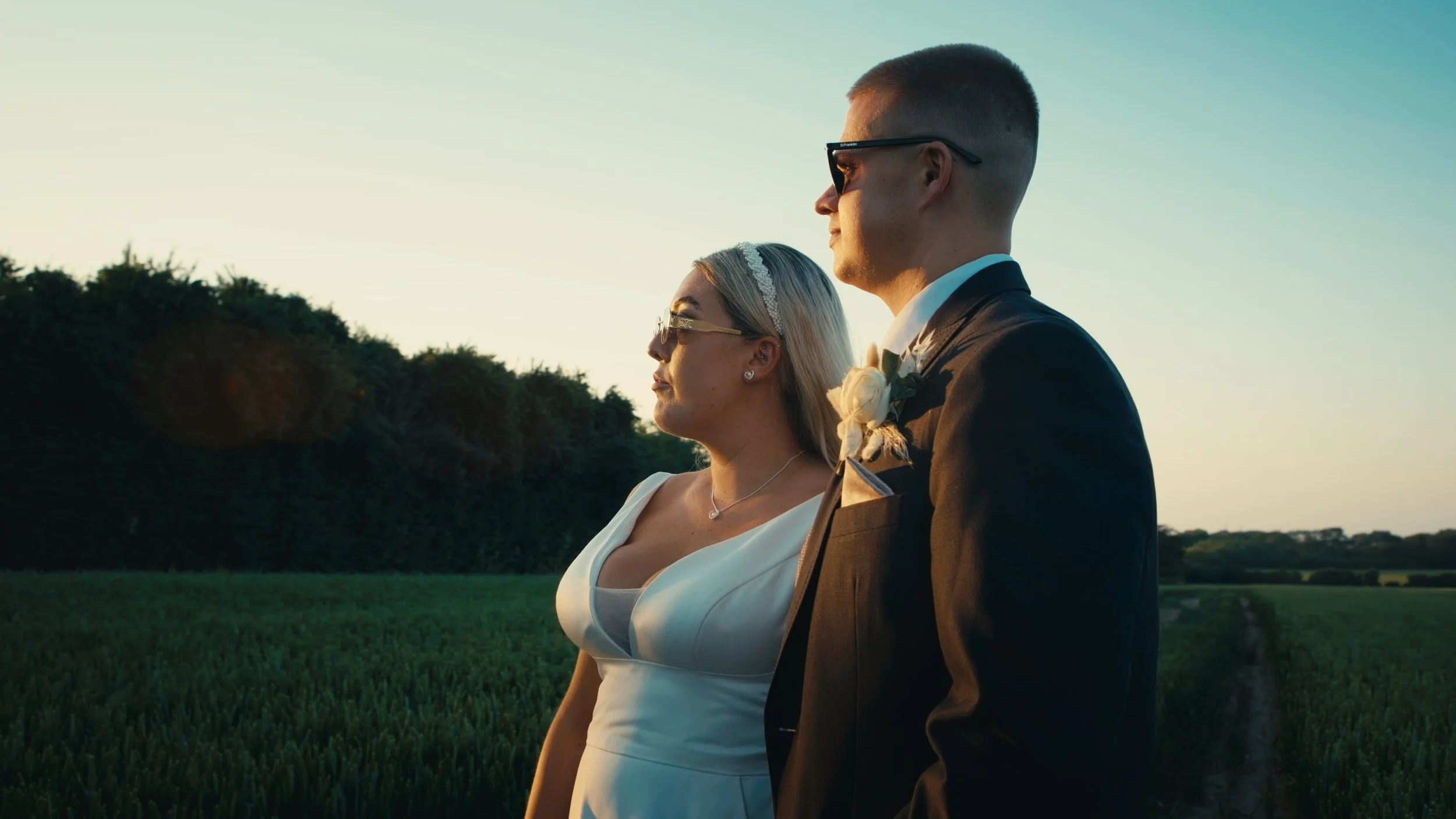 A bride and groom standing outdoors during sunset in a field, facing sideways and wearing formal wedding attire, with trees in the background.