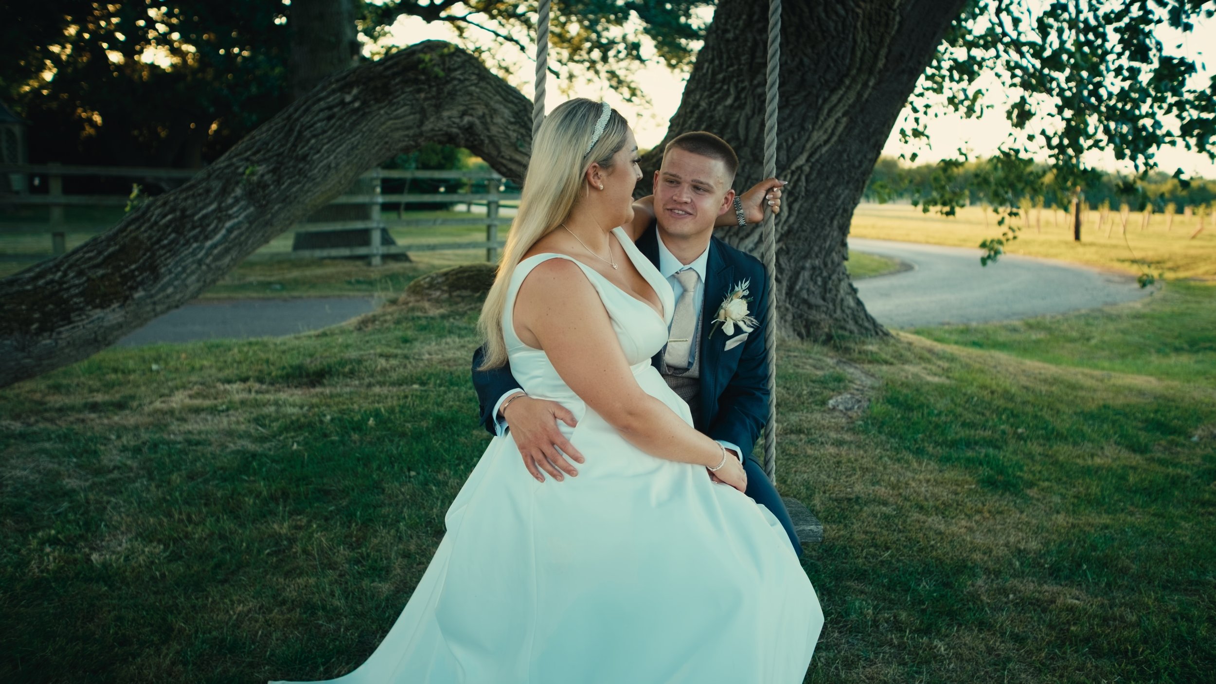 A bride and groom sitting on a swing hanging from a large tree at sunset, with the bride resting her head on the groom's shoulder, both smiling and embracing each other.