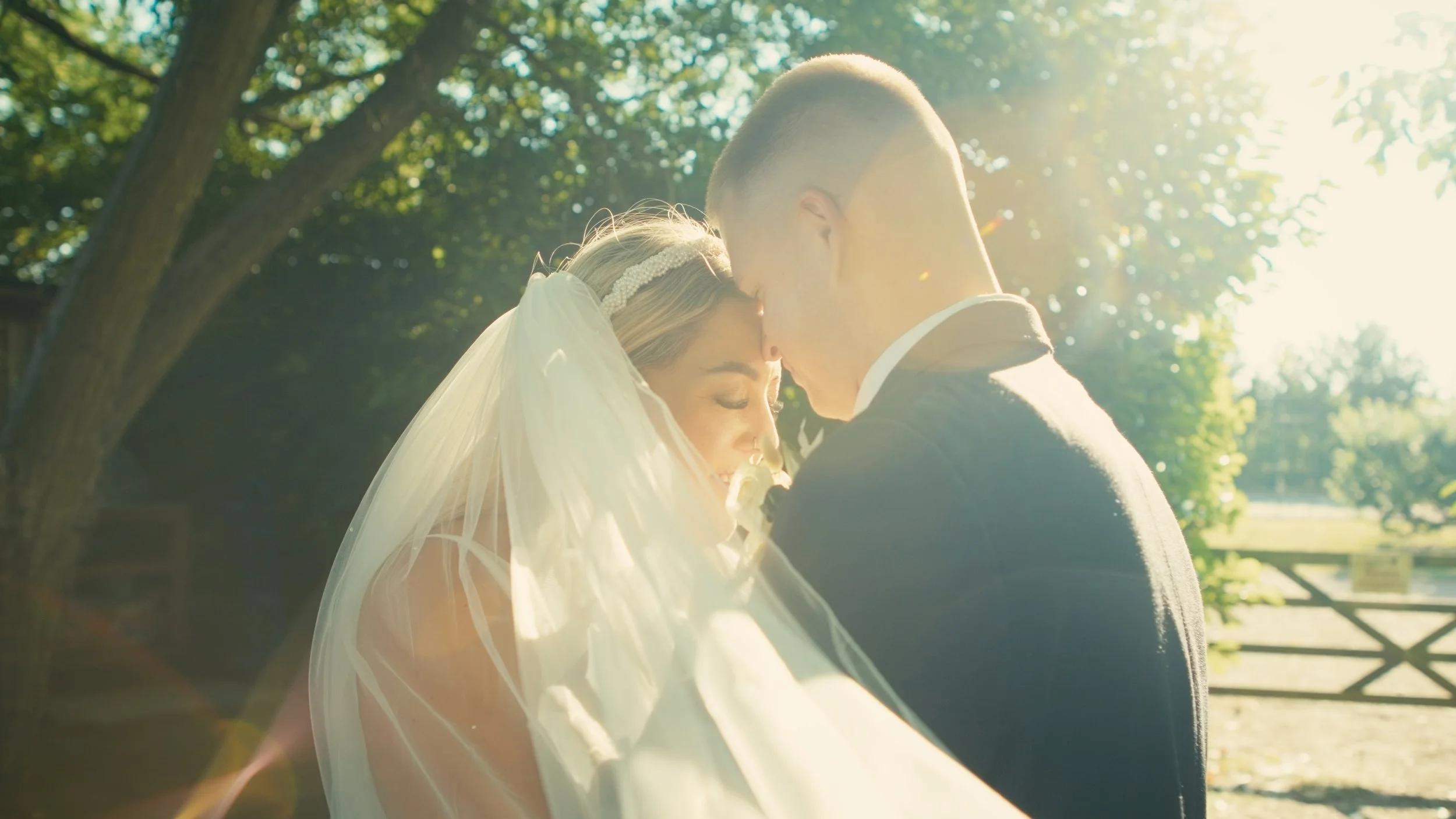 A bride and groom share a tender moment outdoors, with sunlight filtering through trees in the background.