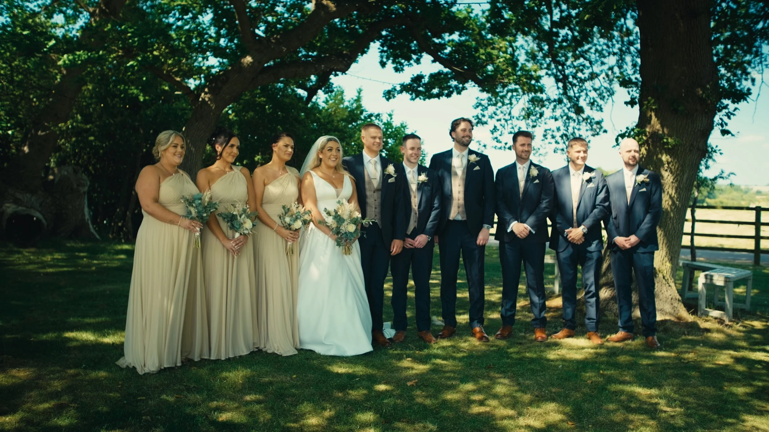 Group of nine people, including a bride in a white wedding dress and six men in dark suits, standing outdoors under large trees during daytime, smiling for a photo at a wedding celebration.