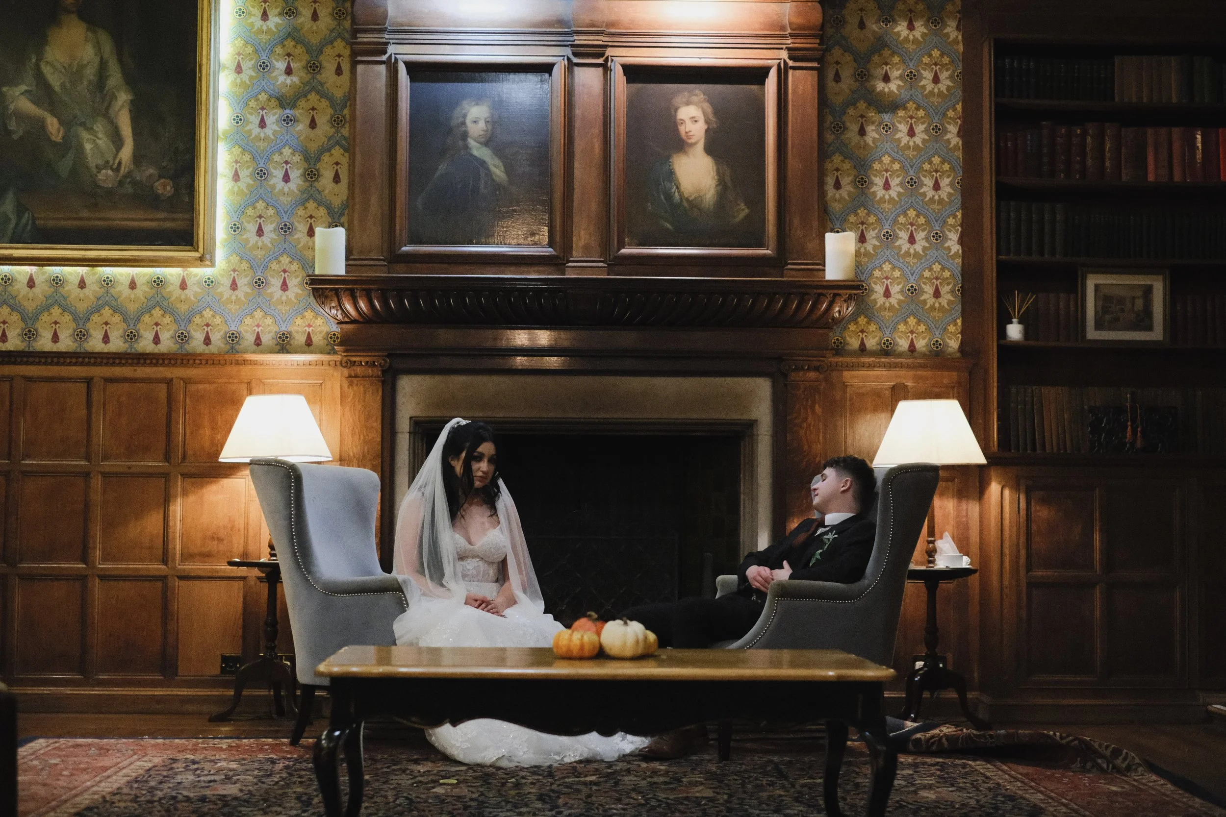 A bride and groom seated in a cozy, wood-paneled room with ornate wallpaper, facing each other. The bride is in a white wedding dress with a veil, and the groom is in a dark suit. There are pumpkins on a table in the foreground, and paintings and can