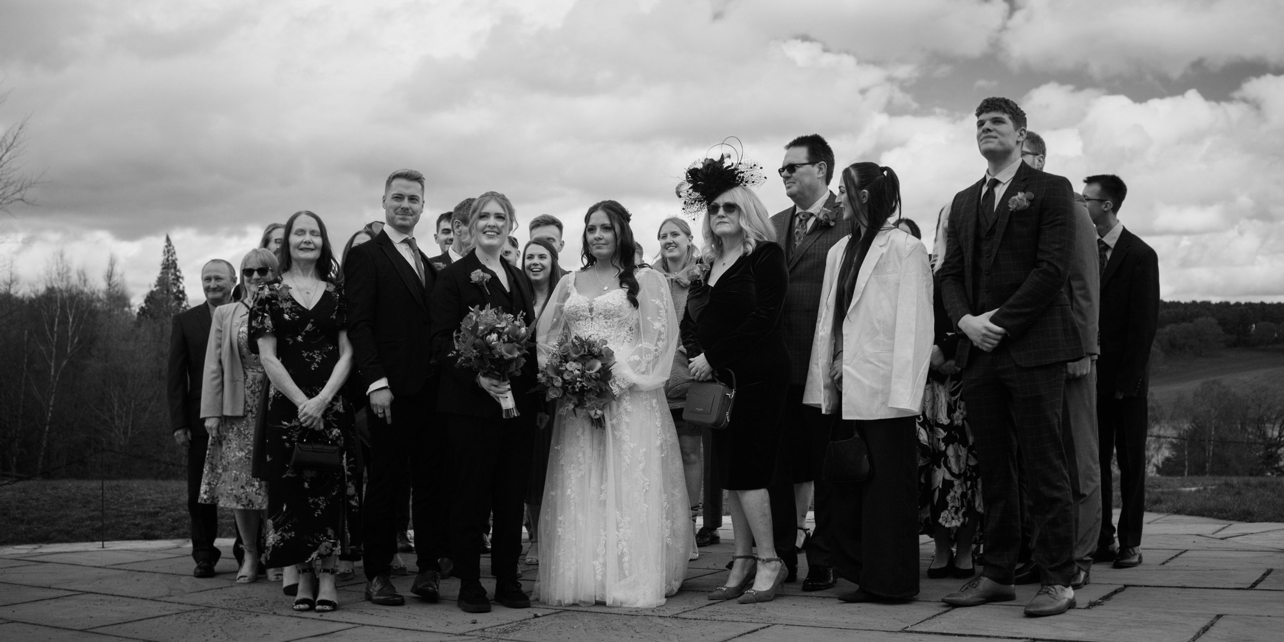 Black and white photo of a wedding group outdoors with cloudy sky, including a bride and groom, family, and friends.