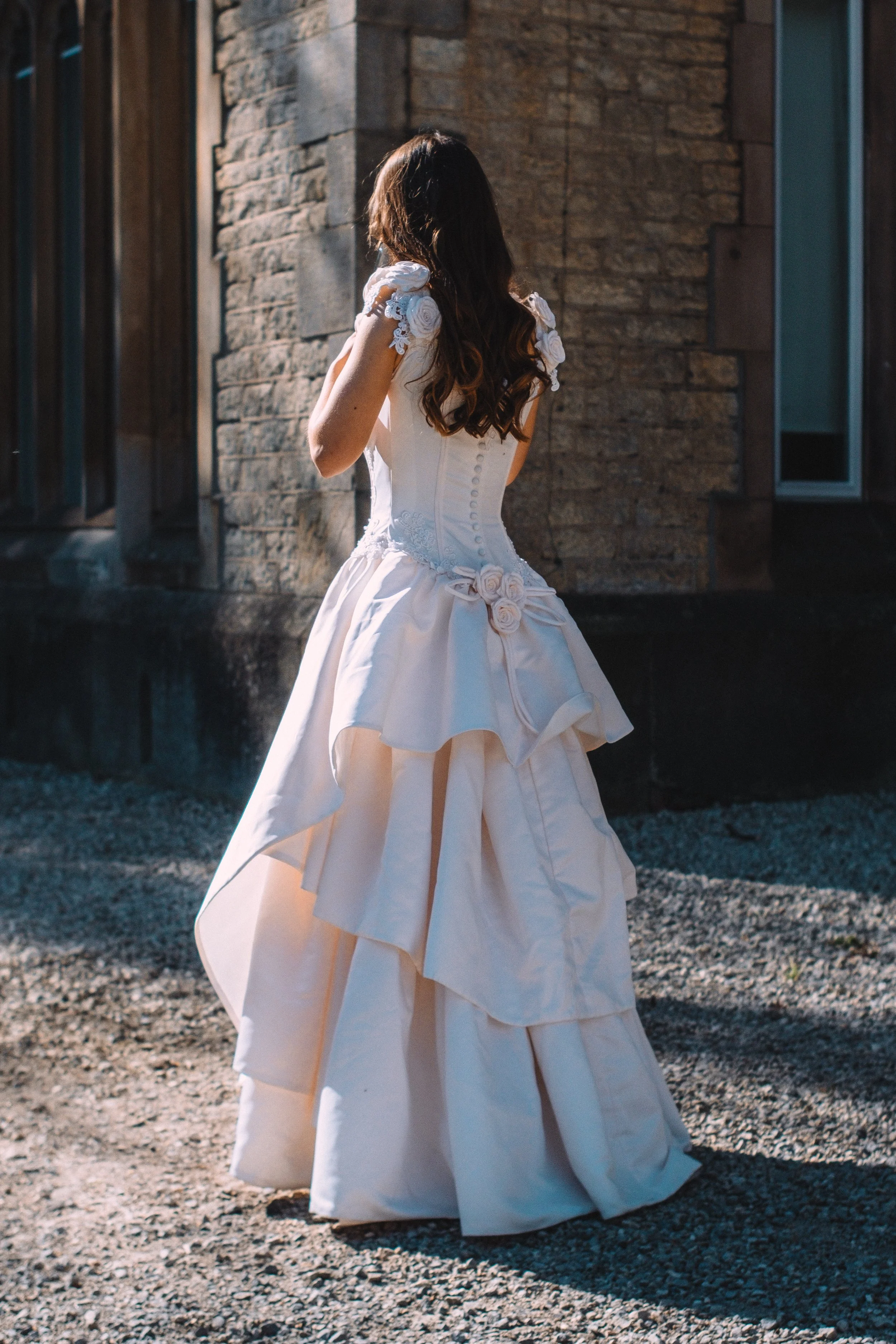 Woman in a white wedding gown with floral details, standing outdoors near a stone wall, with her back to the camera.