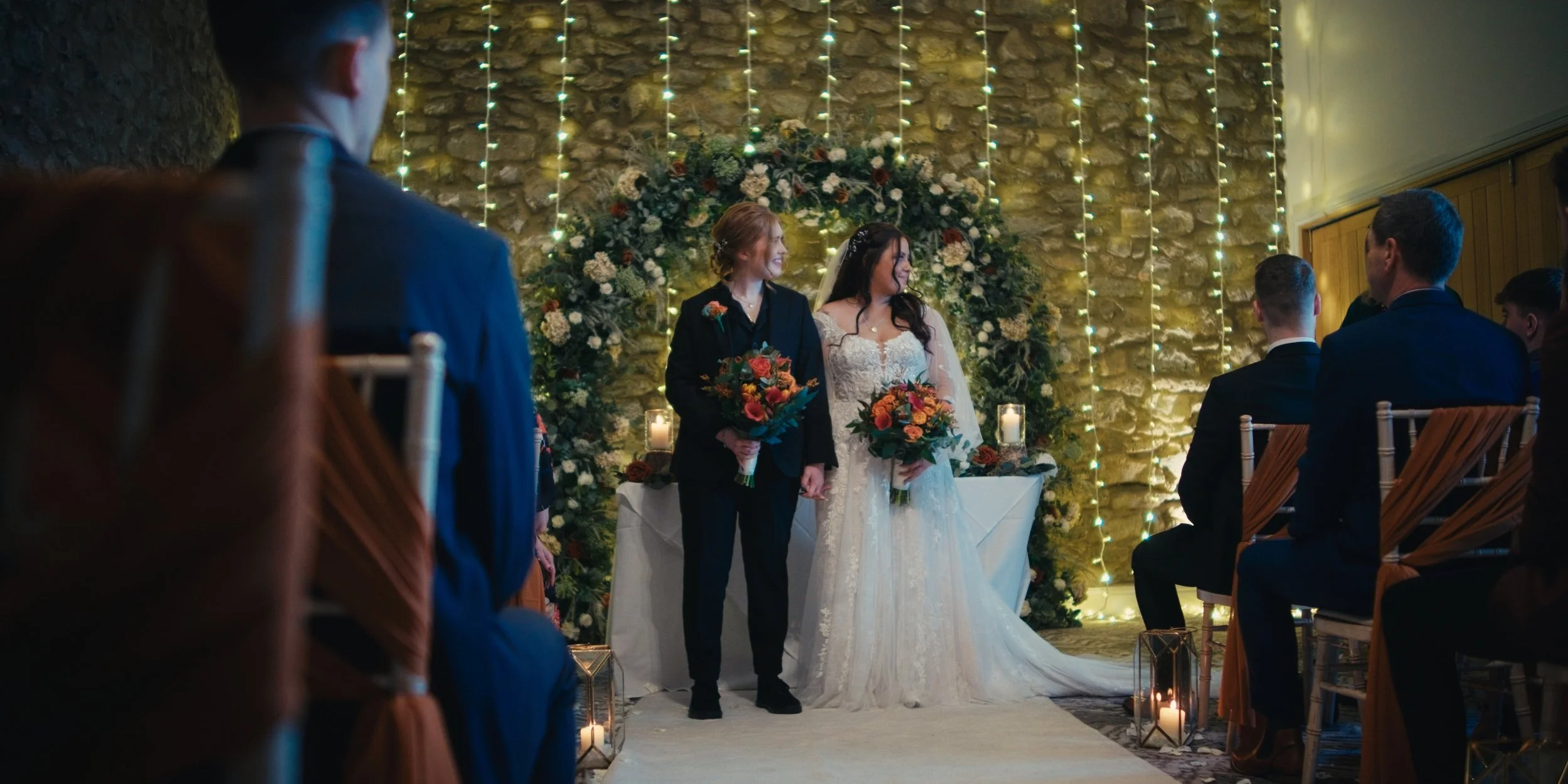 A wedding ceremony with two women holding bouquets, standing in front of a stone wall decorated with string lights and floral arrangements, surrounded by seated guests.