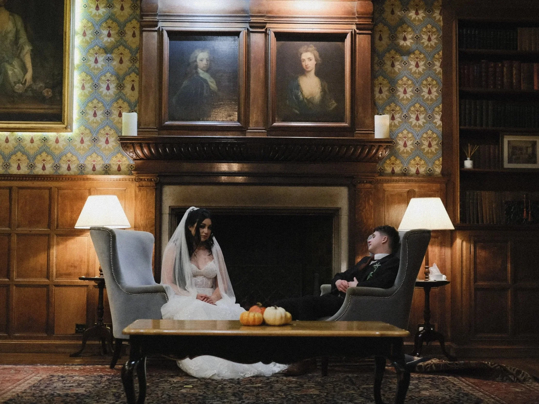 A wedding ceremony taking place in an elegant wooden-paneled room with paintings on the wall. The bride, wearing a white wedding dress and veil, and the groom, dressed in a formal black tuxedo, are seated facing each other, holding hands. A small table with pumpkins is in the foreground.