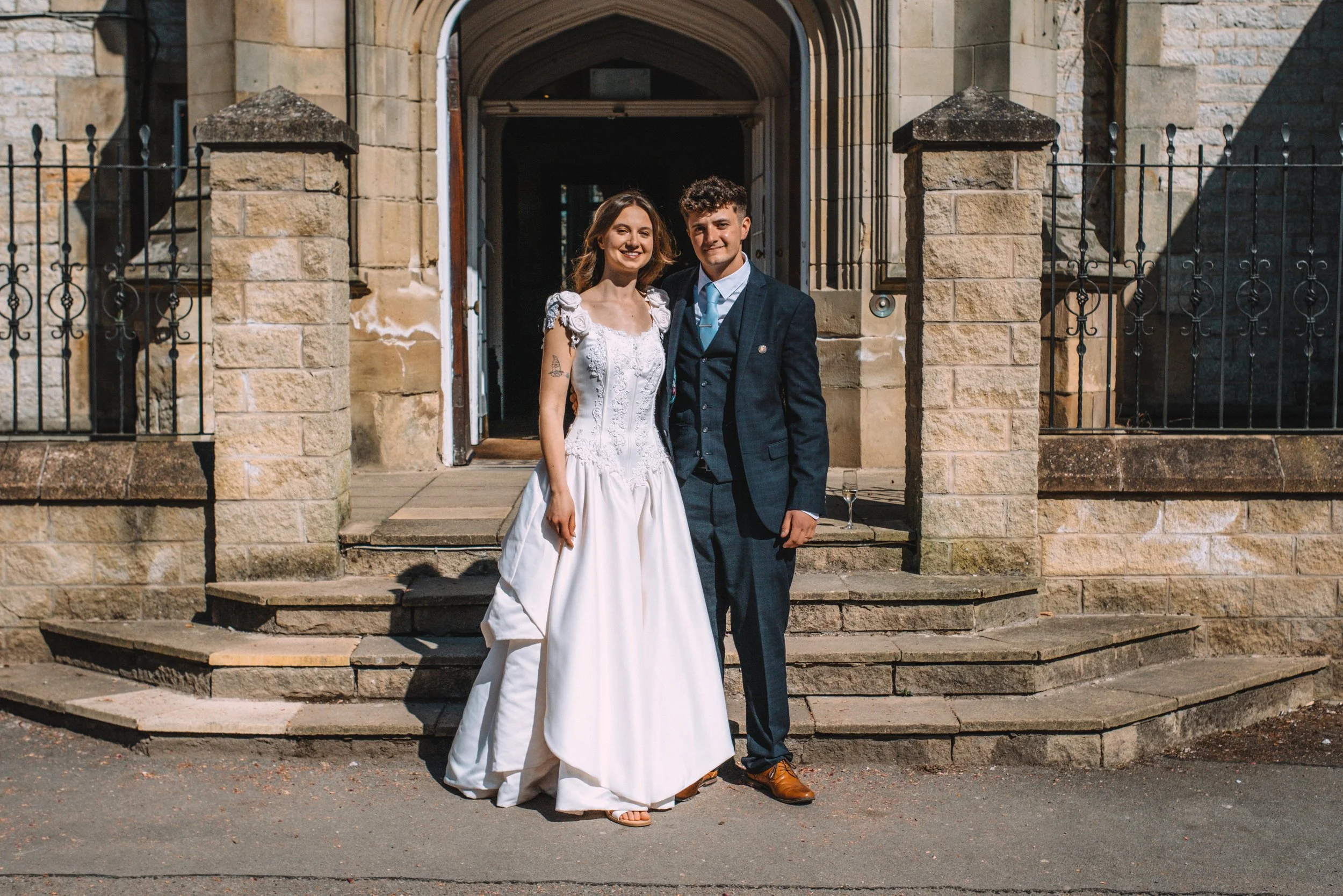 A bride and groom standing on the steps outside a stone church, smiling for a photo, with the bride in a white wedding dress and the groom in a dark suit.