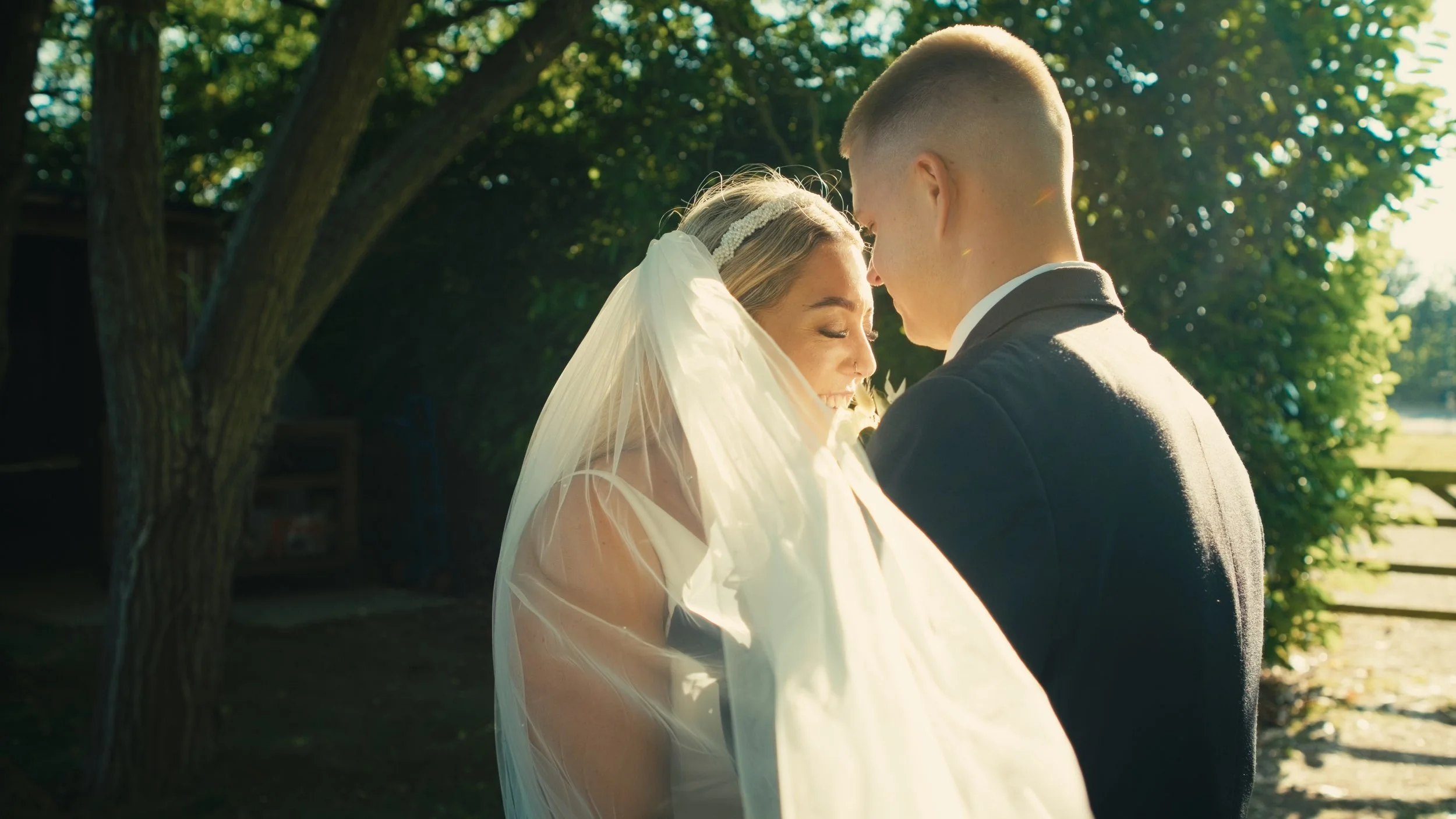 Bride and groom embrace outdoors in sunlight, smiling with foreheads touching under a tree.