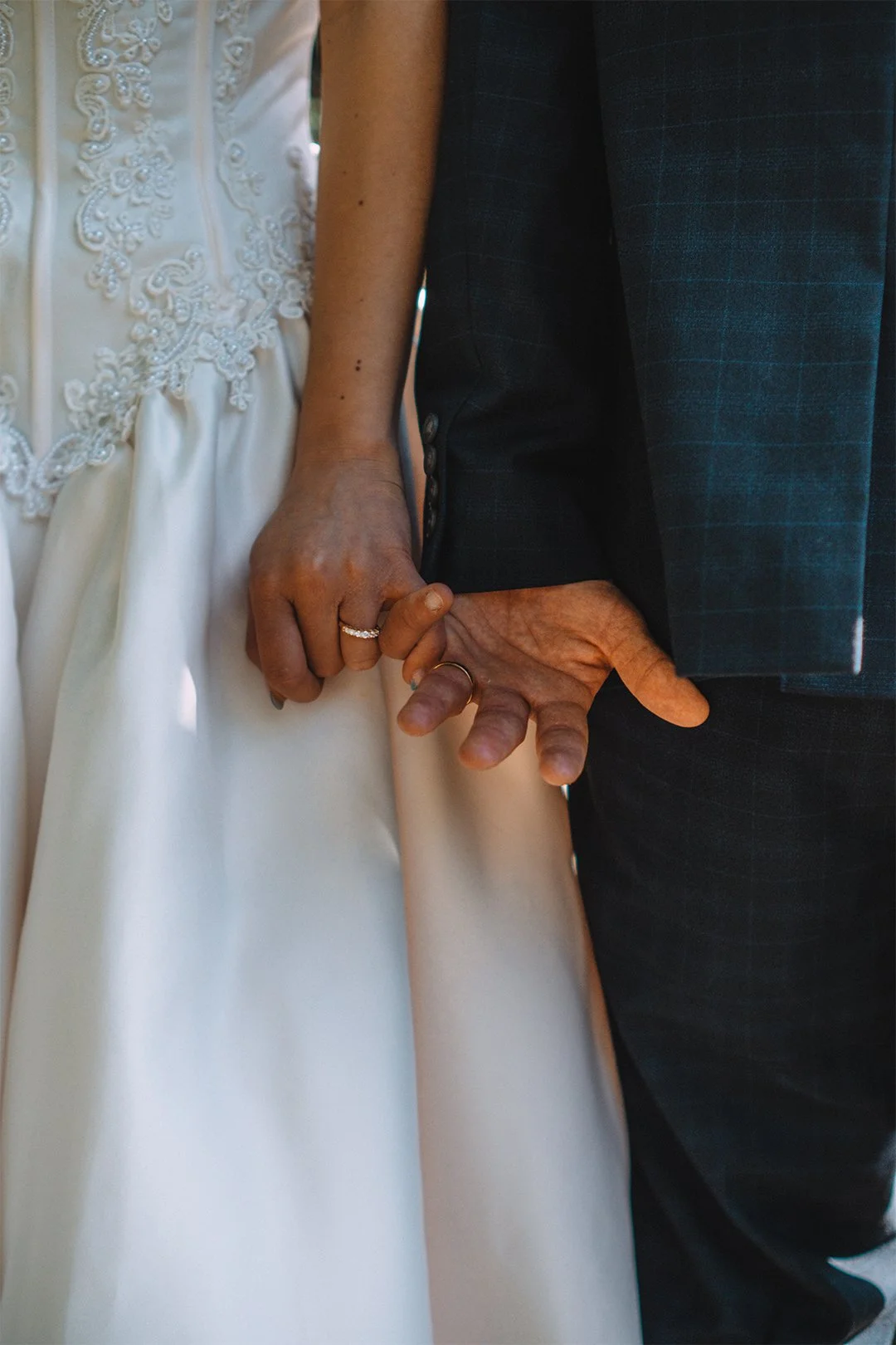 Close-up of a couple holding hands, with the woman's hand resting on the man's hand. The woman is wearing a wedding dress with embroidery, and the man is dressed in a dark suit. Both are wearing rings.