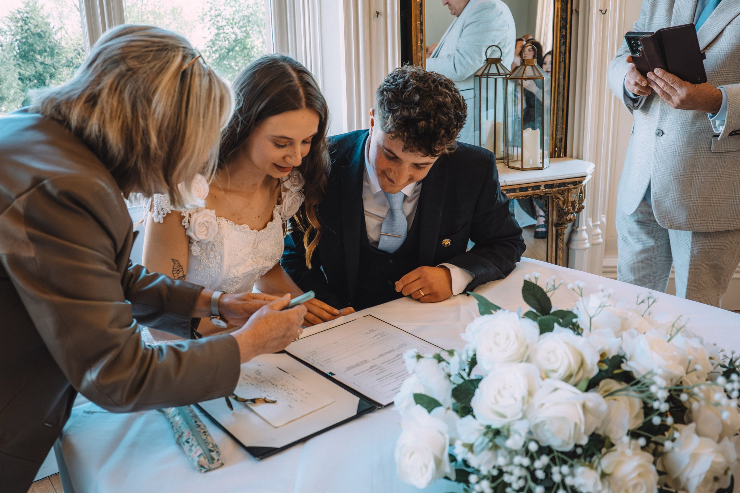 People signing documents at a wedding reception table decorated with white flowers, with a window showing trees in the background.