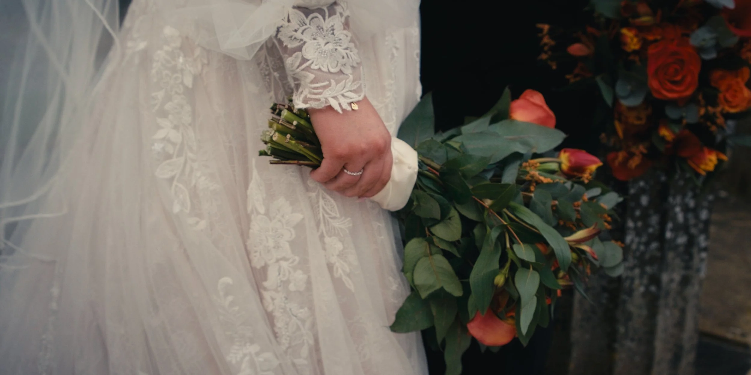 Close-up of a bride's hand resting on a white lace wedding dress, holding a bouquet of green foliage and orange flowers.