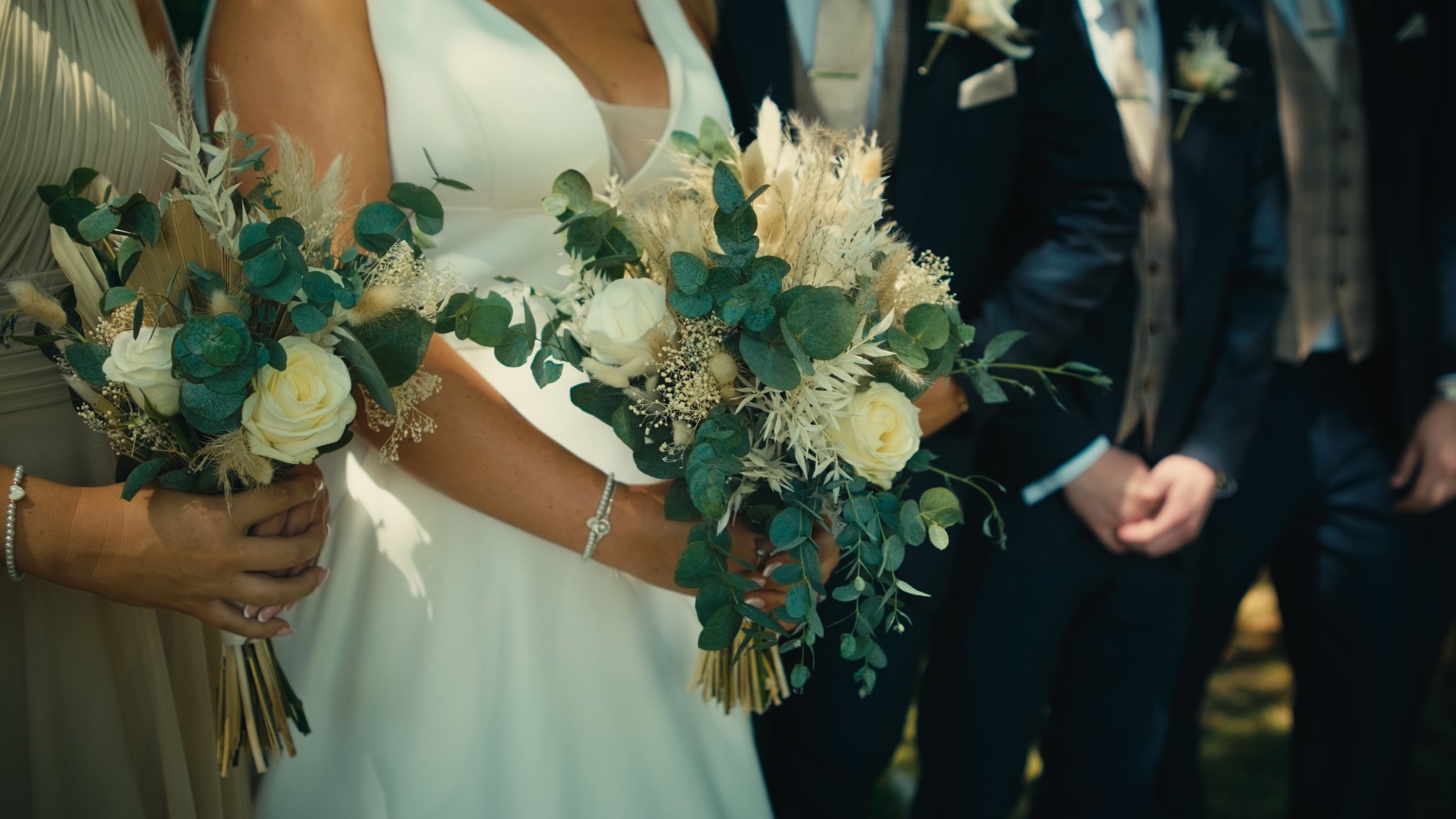 A bride holding a bouquet of white roses, green foliage, and dried flowers during a wedding ceremony, standing next to a person in a beige dress, with a man in a dark suit and light-colored shirt standing nearby.