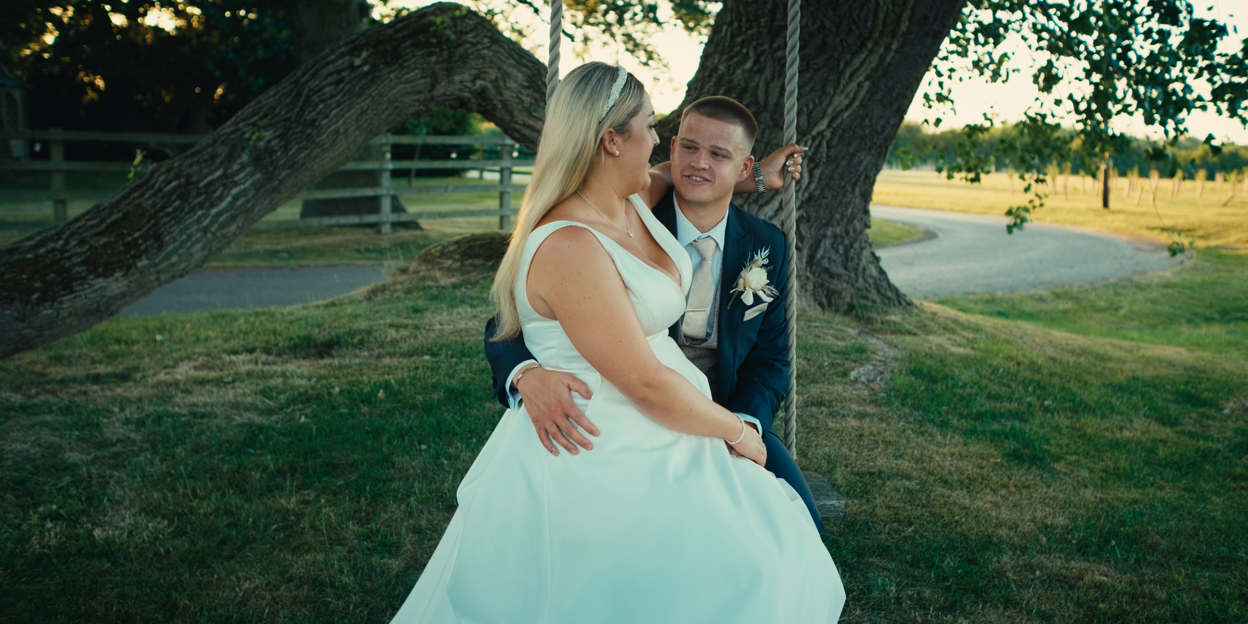 A bride and groom sitting on a swing under a large tree in a park with green grass and a winding path.