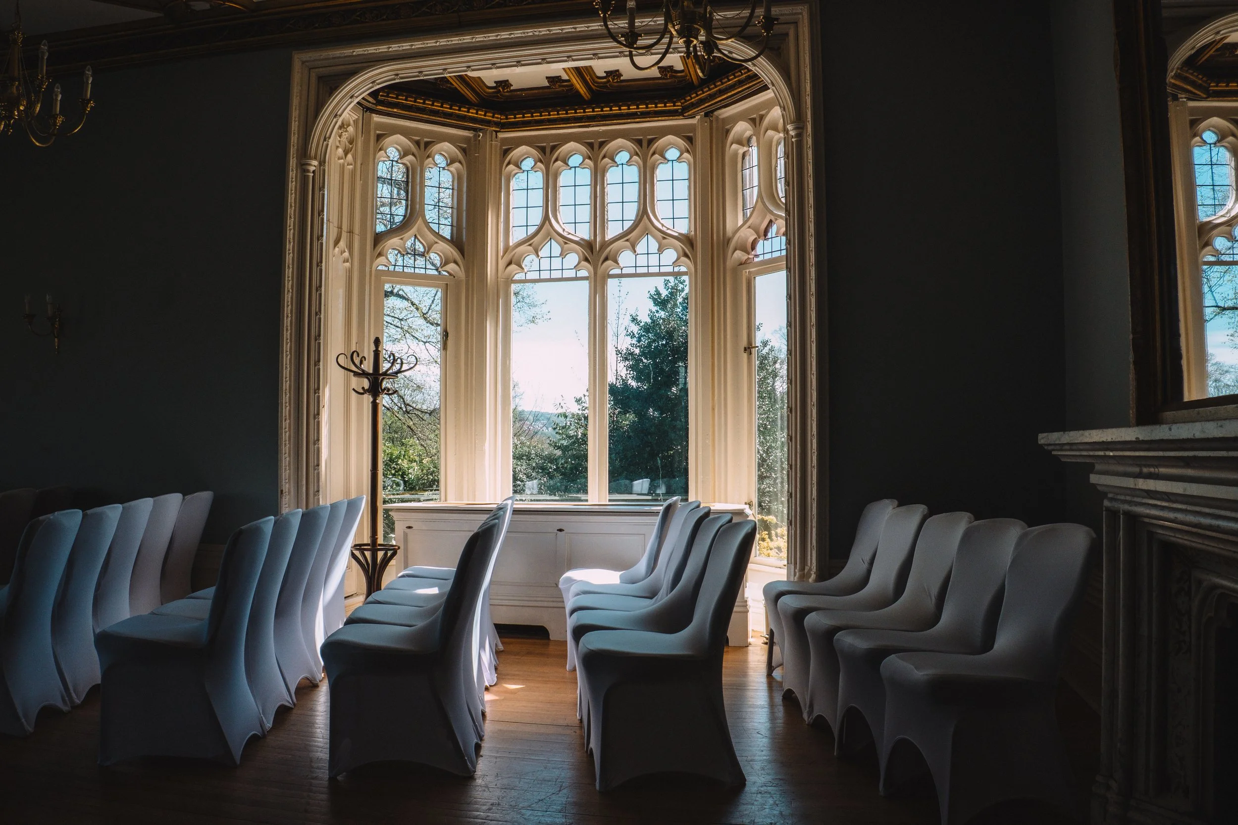 Elegant banquet room with white chairs arranged around a long table, large ornate window with Gothic-style arches letting in natural light, dark walls, and a chandelier hanging from the ceiling.
