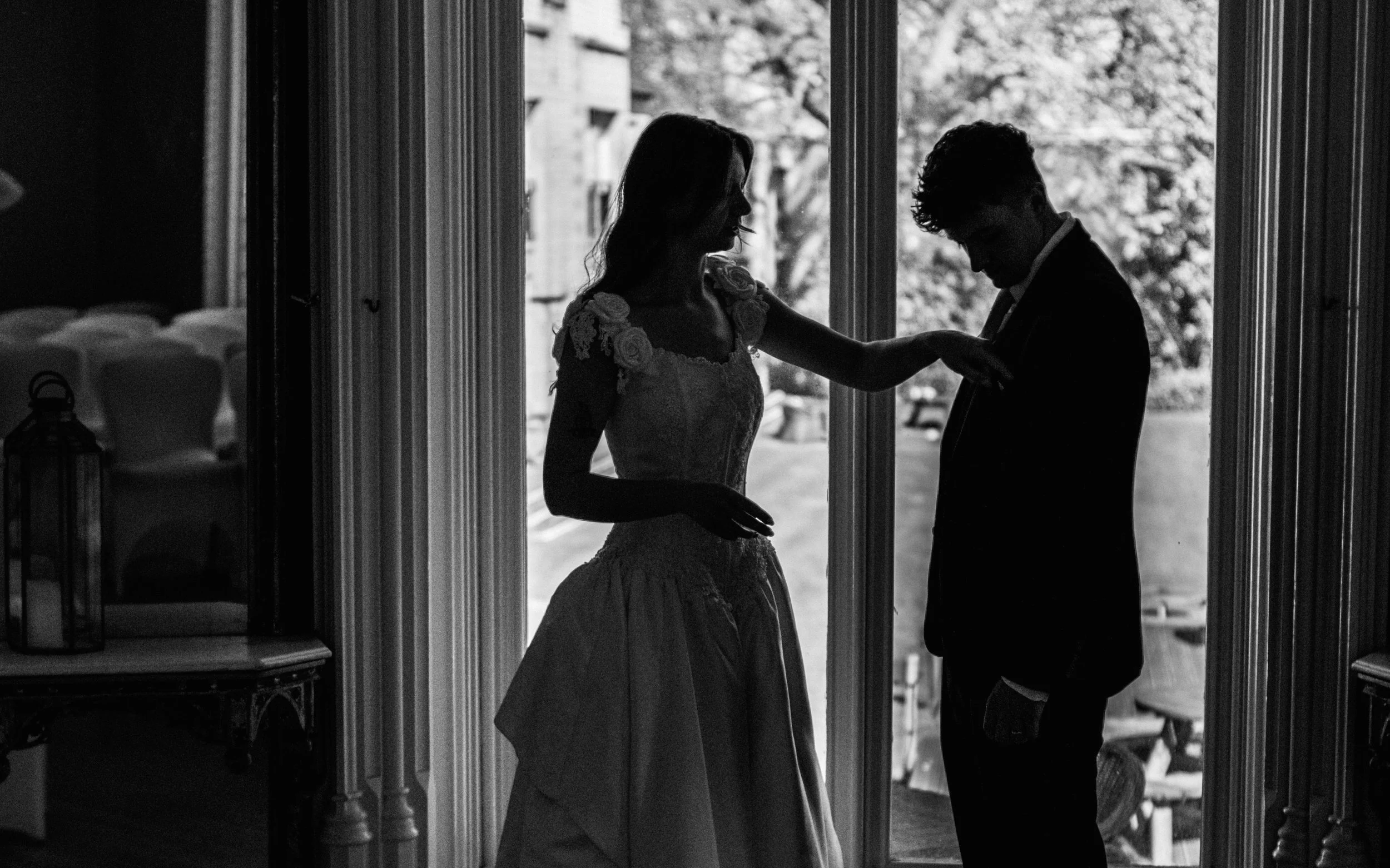 Silhouetted bride and groom inside near a large window, with trees visible outside, during a wedding.