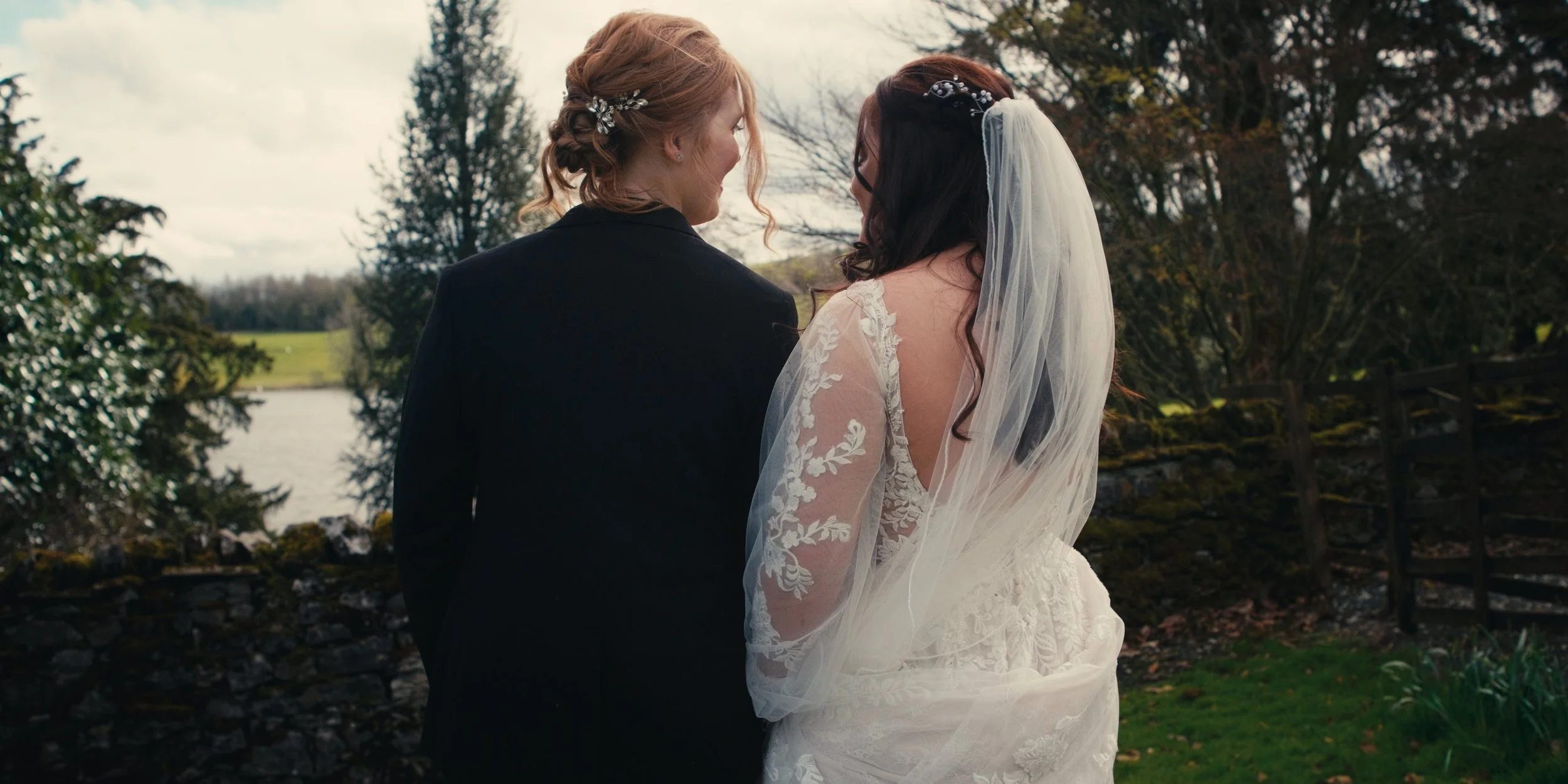 A bride and groom standing outdoors near a lake, talking and smiling at each other, with trees and a stone fence in the background.