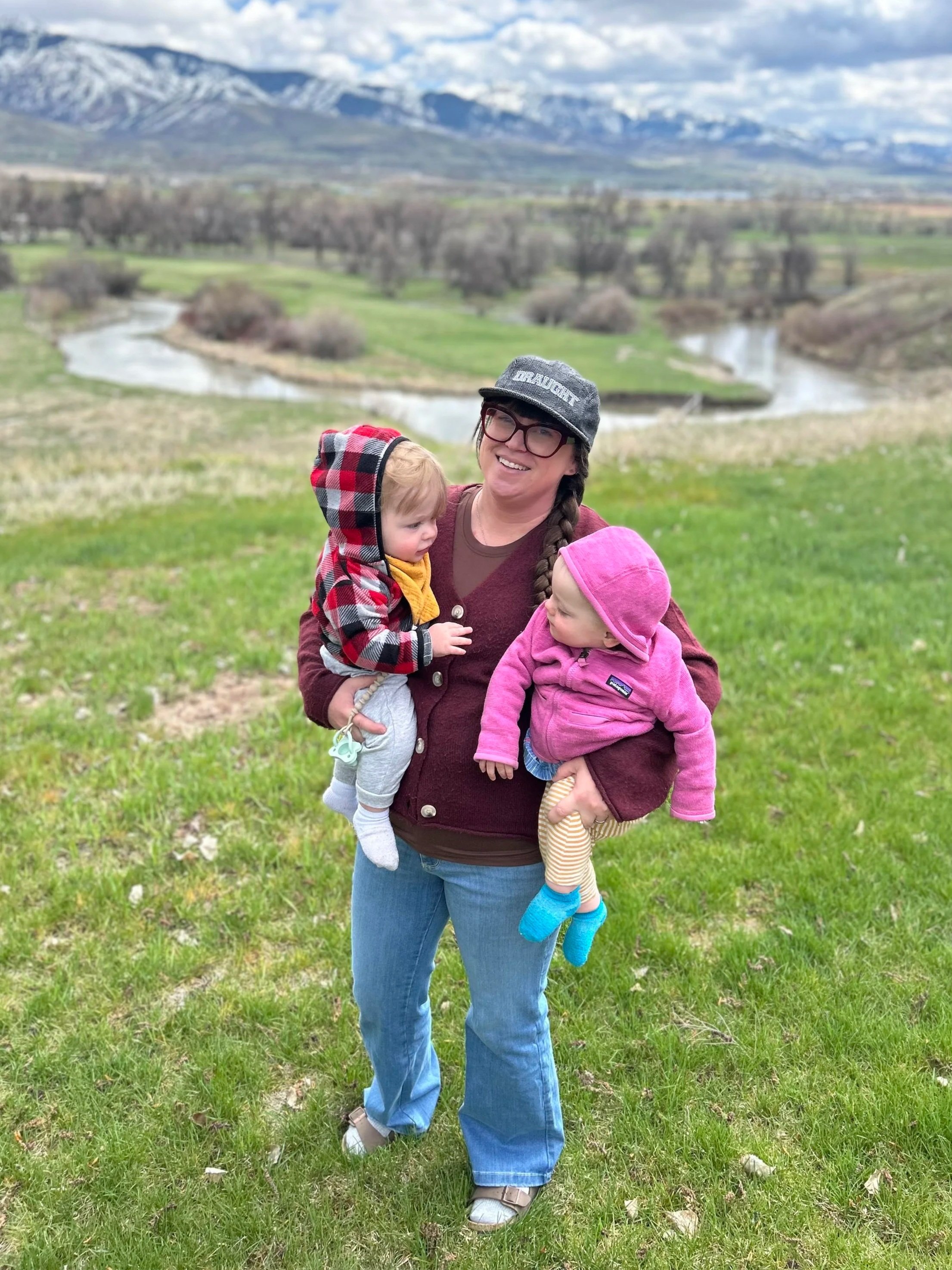 A woman with glasses, wearing a gray cap with 'DRAUGHT' written on it, holding two young children outdoors in a grassy field, with mountains, a river, and a cloudy sky in the background.