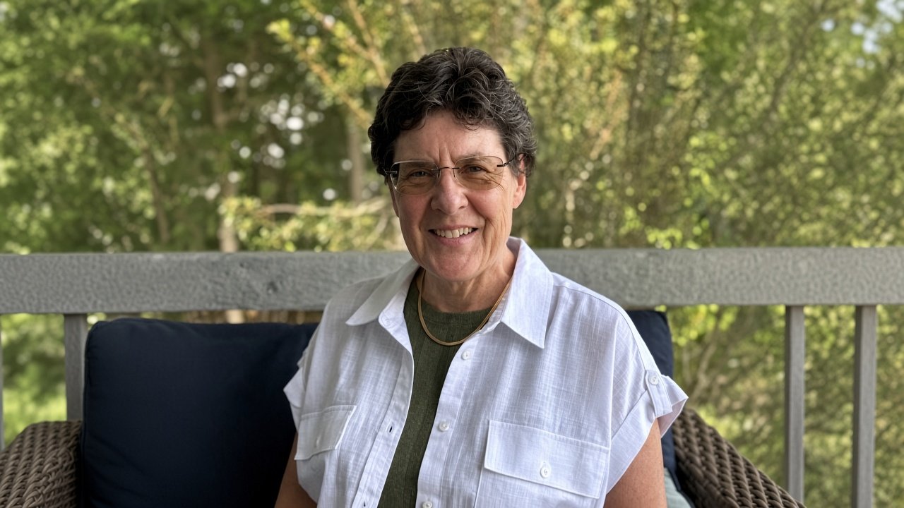 Smiling woman with short dark hair, glasses, and a white shirt sitting outdoors on a patio with green trees in the background.