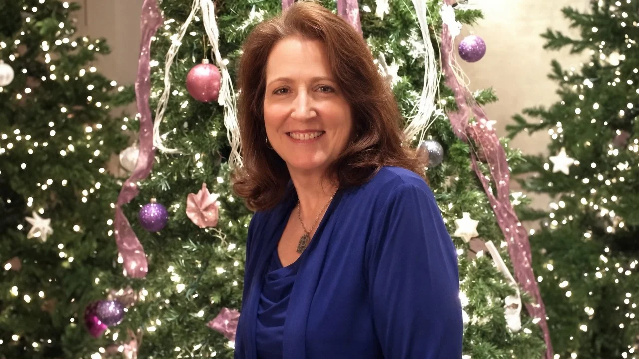 A woman with brown hair smiling and wearing a blue dress, standing in front of decorated Christmas trees with pink, purple, and white ornaments and ribbons, lit with string lights.