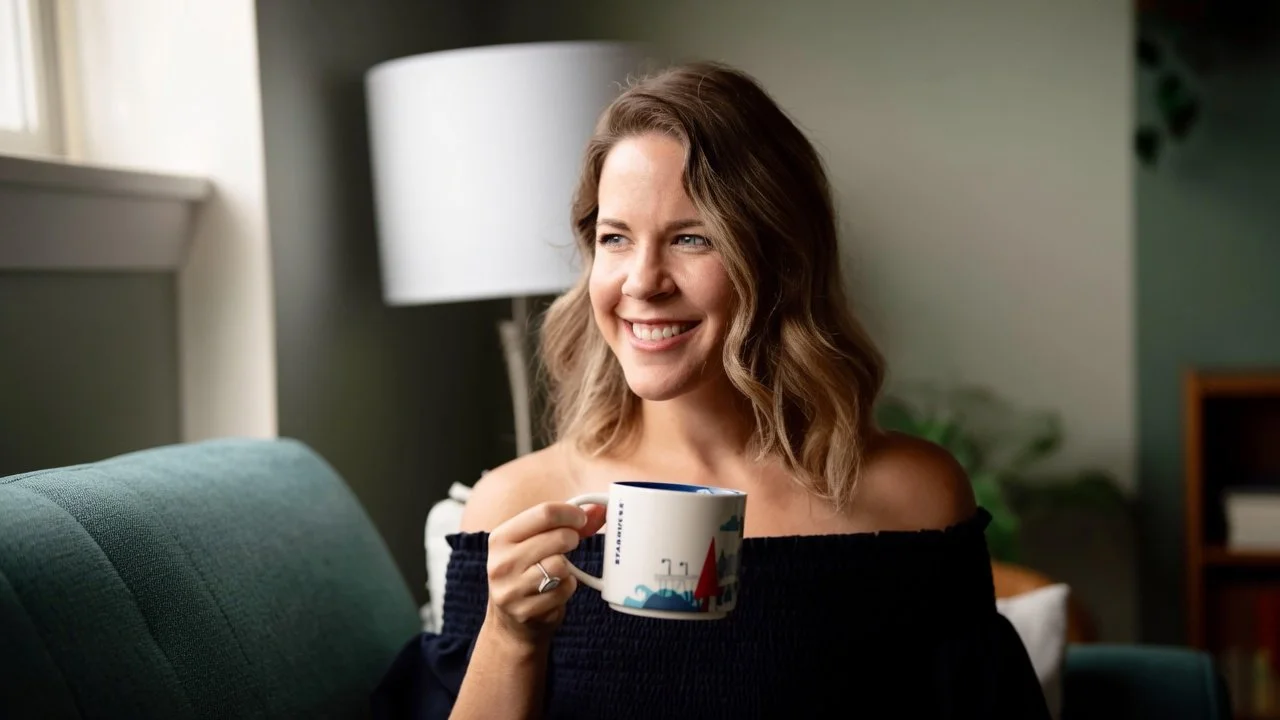 A woman with shoulder-length wavy hair smiling while holding a Starbucks mug in her right hand, sitting on a teal couch in a cozy living room.