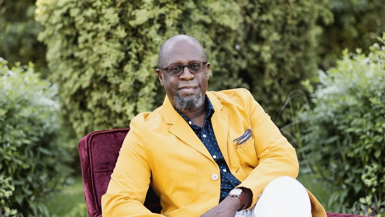A man with glasses, a beard, and a bald head sitting outdoors on a red chair, wearing a yellow blazer and a dark blue shirt with white polka dots, with greenery in the background.