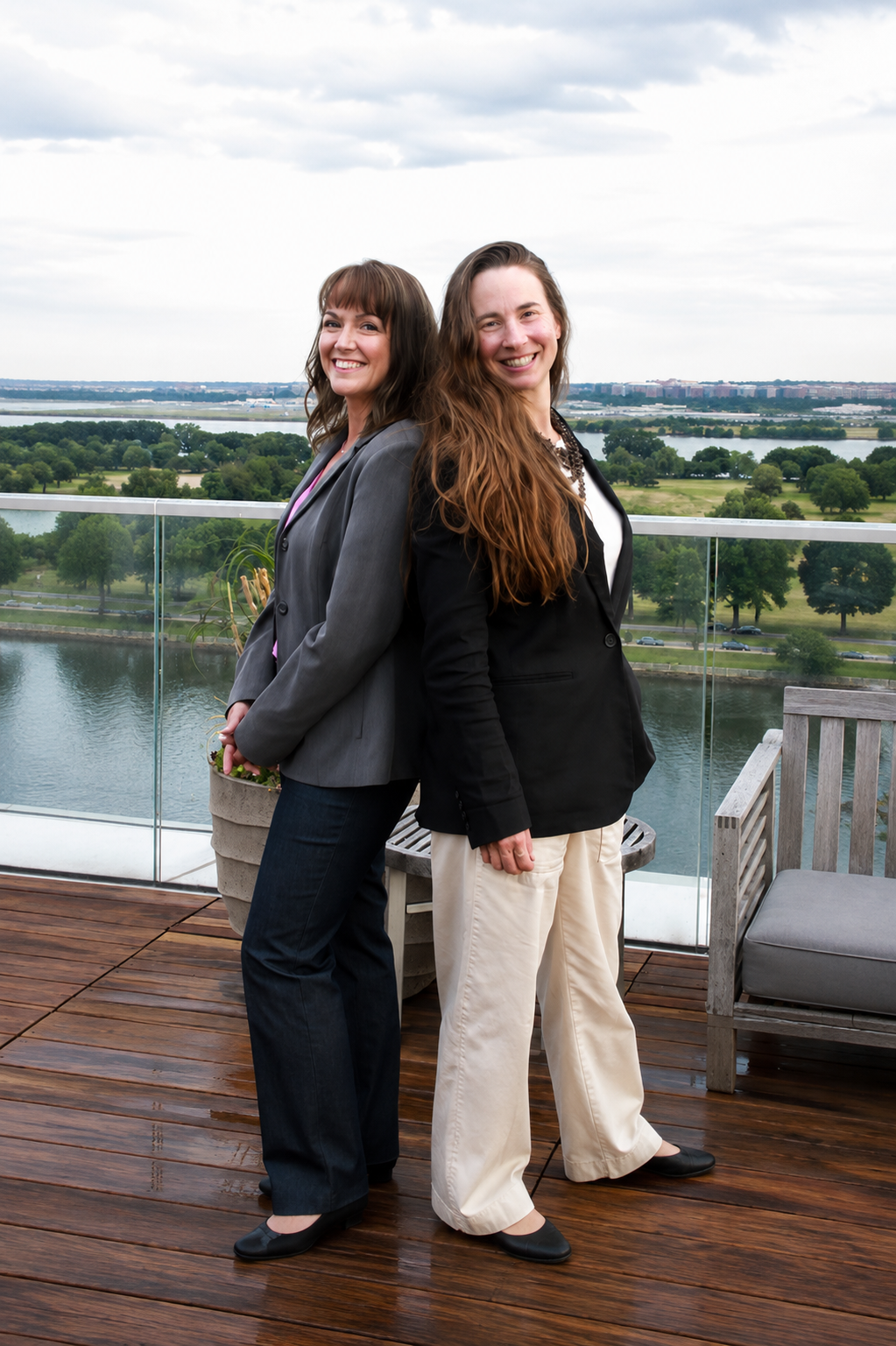 Two women stand back to back on a rooftop deck overlooking a river and green landscape with trees and a cloudy sky, smiling at the camera.