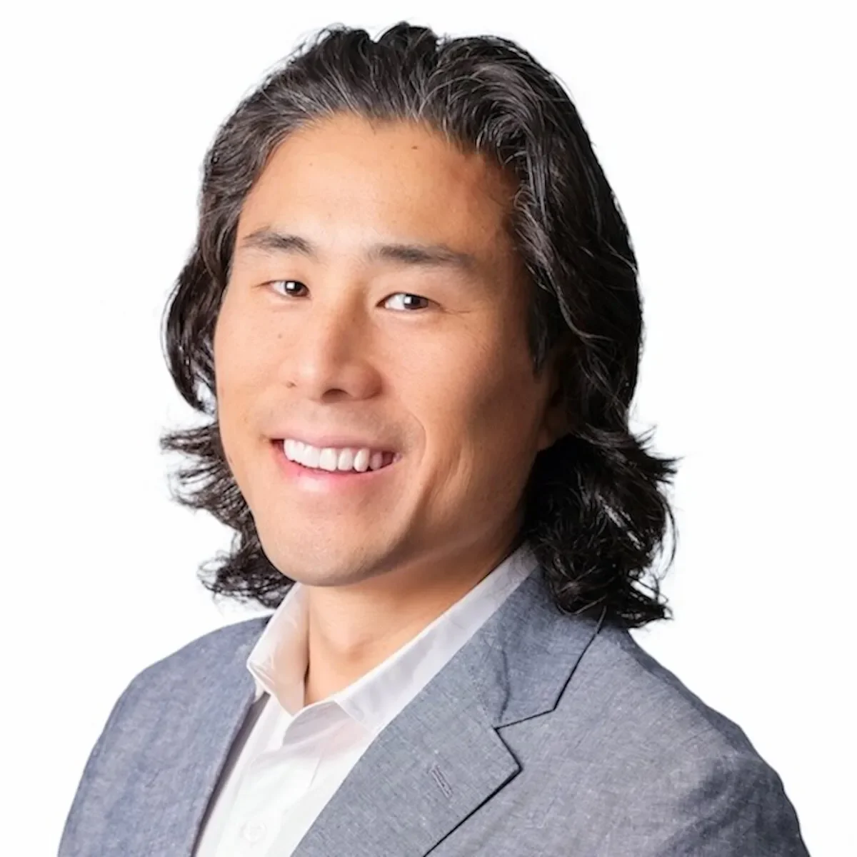 A portrait of a man with long, wavy black hair, wearing a light gray suit jacket and a white shirt, smiling against a plain white background.