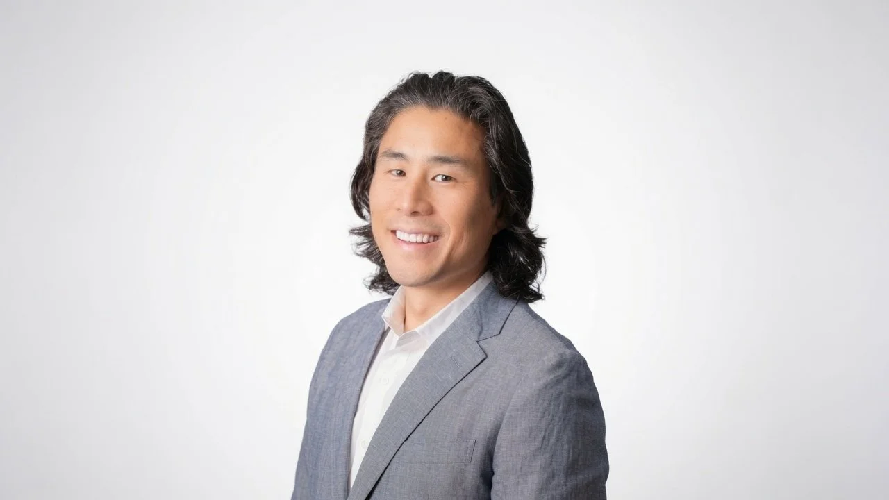 A man with long black hair smiling, wearing a gray blazer and white shirt, in front of a plain light gray background.