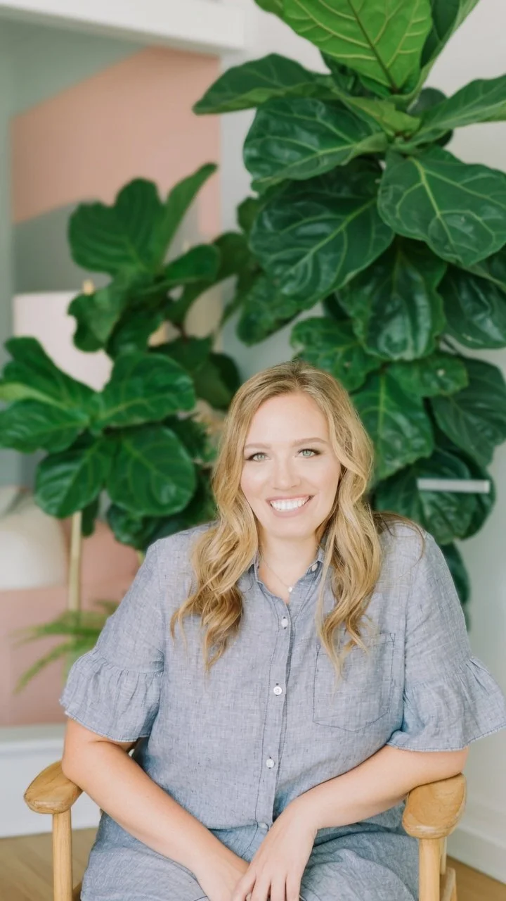 A woman with blonde hair smiling and sitting in a wooden chair in front of large green houseplants.